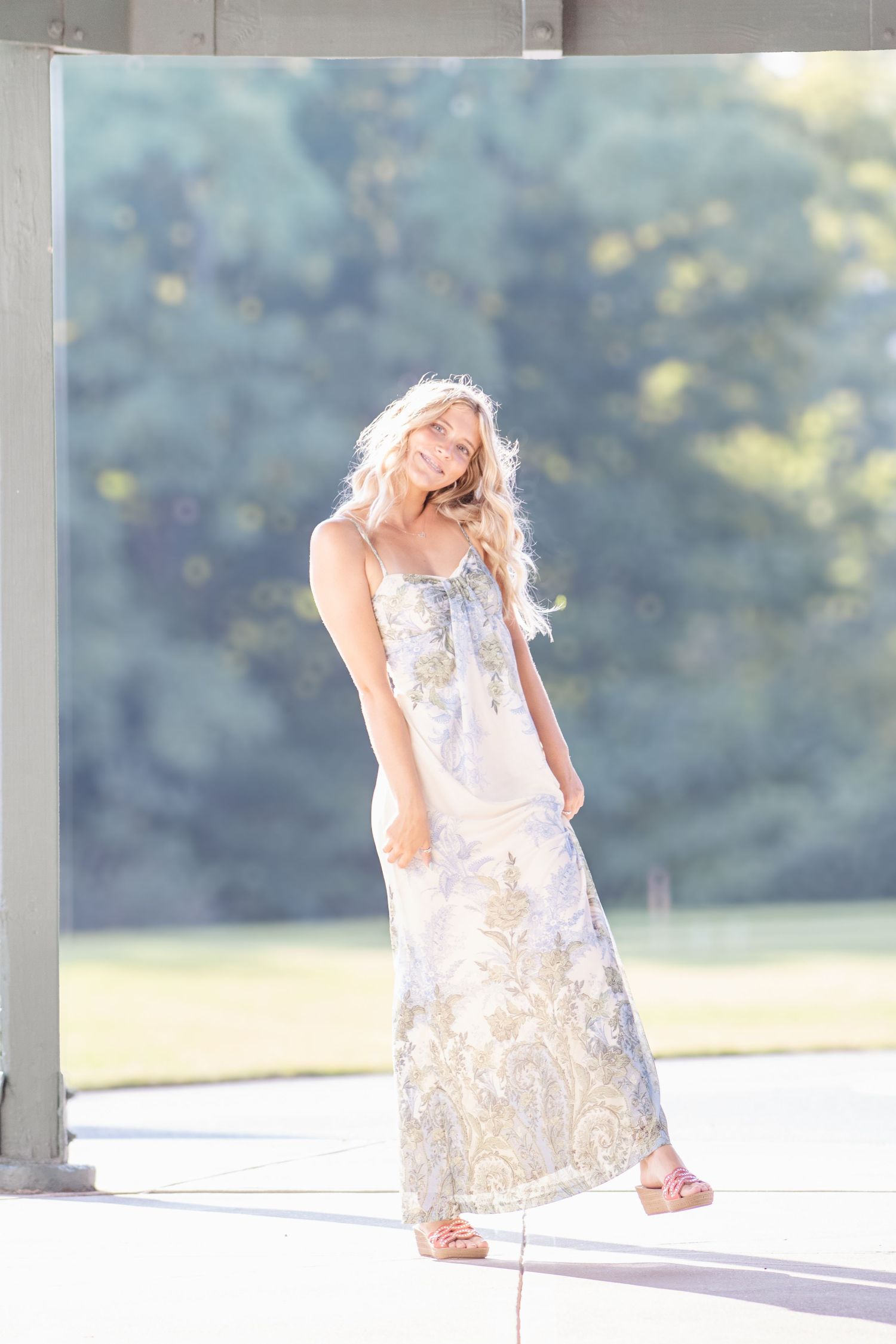 Blonde woman in floral maxi dress standing in sunlit outdoor space with lush green trees in background.