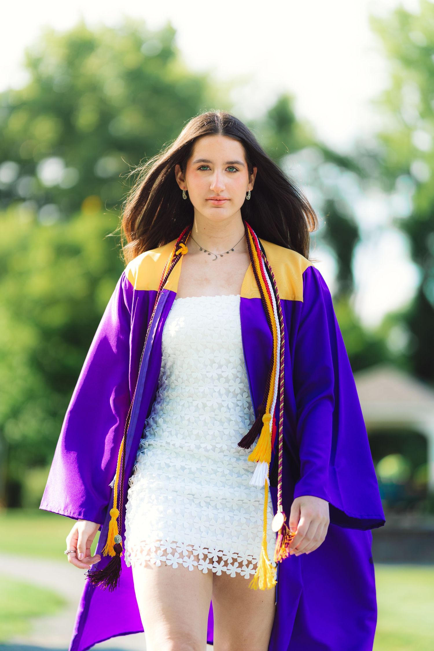 Young woman in purple and gold graduation gown walking outdoors on a sunny day.