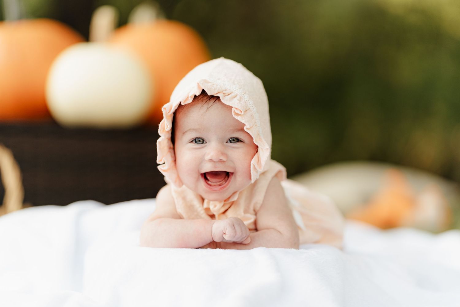 Smiling baby in a pink bonnet lying on a white blanket surrounded by pumpkins in a fall outdoor setting.