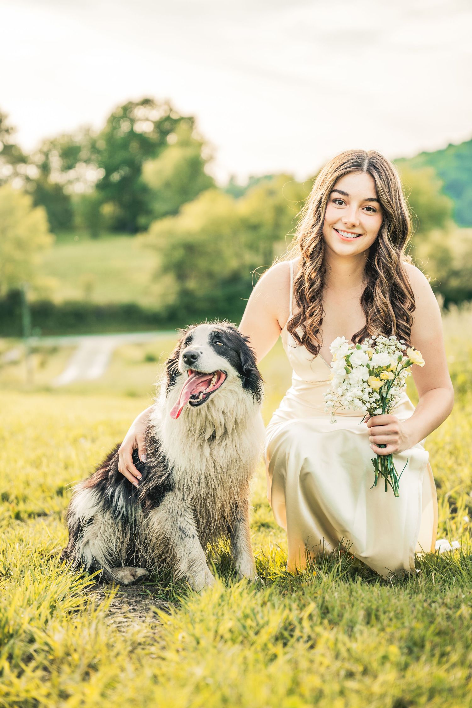 Young woman in white dress holding wildflowers poses with border collie dog on green grass outdoors.