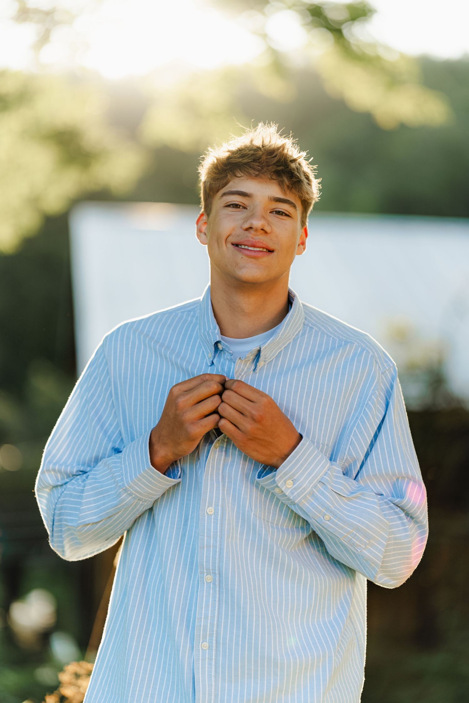 Smiling teenage boy in a light blue striped button-up shirt posing outdoors in golden sunlight.