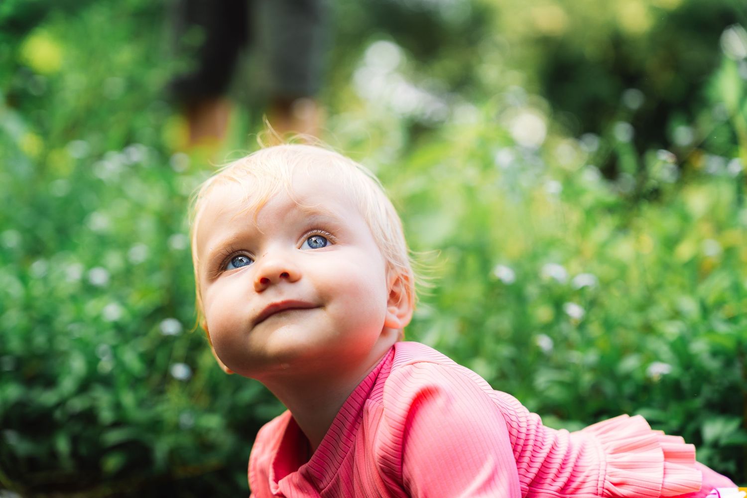 Adorable blue-eyed baby in pink outfit looking up curiously in a lush garden.