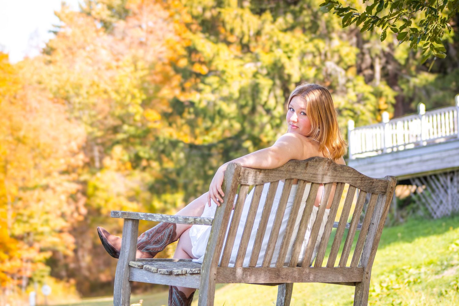 Woman in floral dress sitting on wooden park bench surrounded by vibrant autumn foliage on a sunny day.
