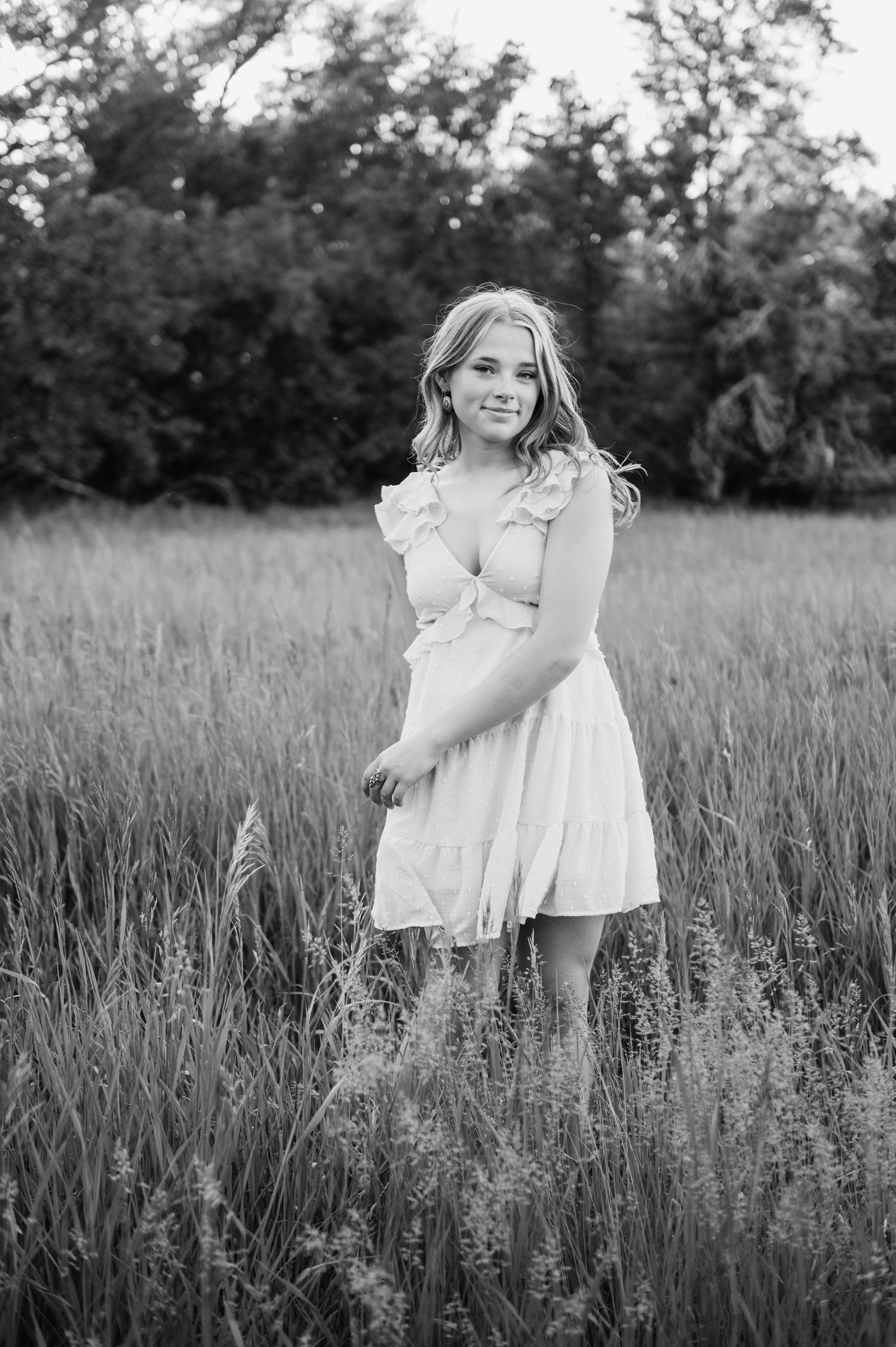 Black and white portrait of a young blonde woman in a white dress standing in a tall grass field.