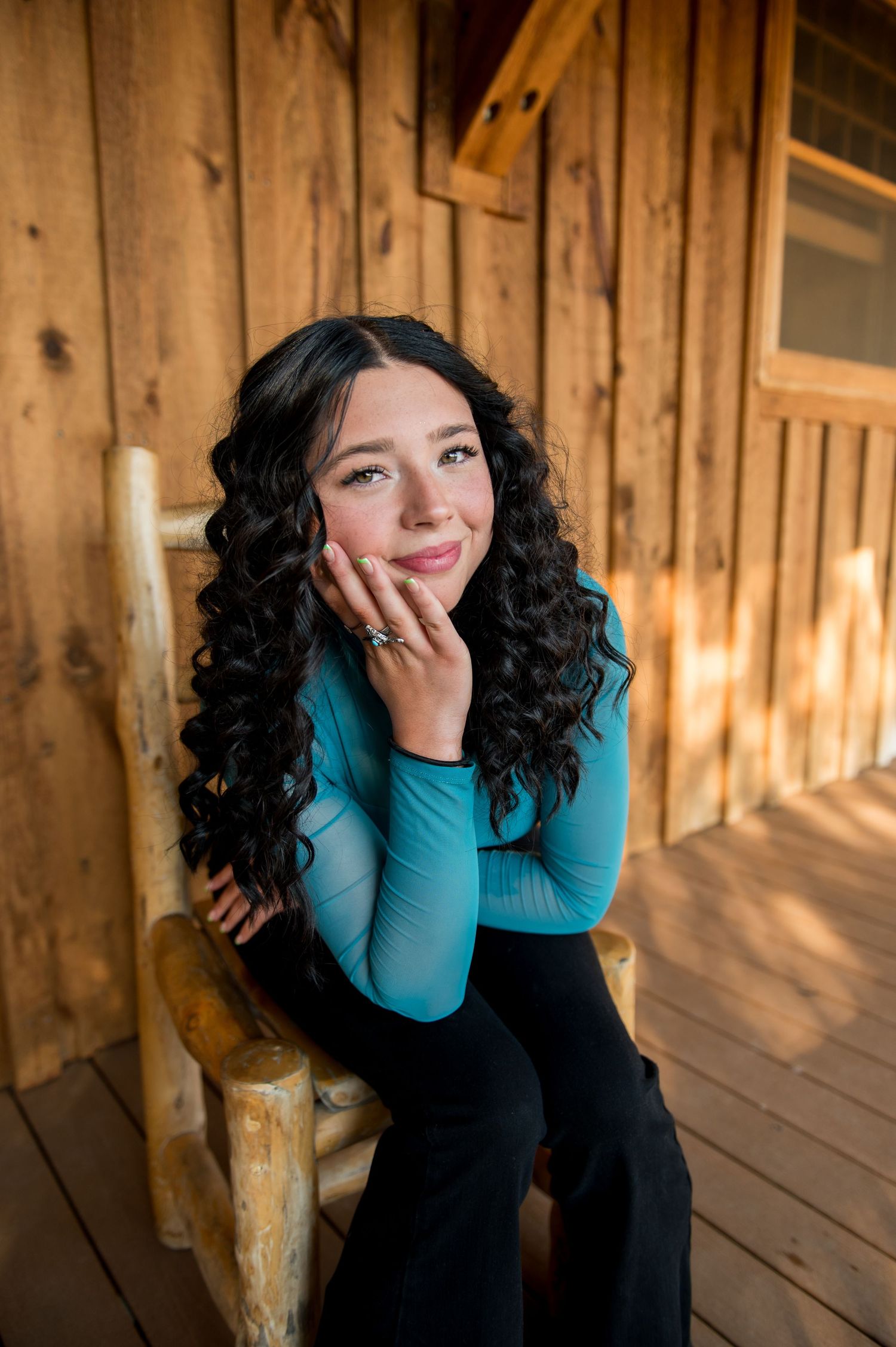 Young woman with curly black hair in a teal top sitting on a rustic log chair on a wooden porch.