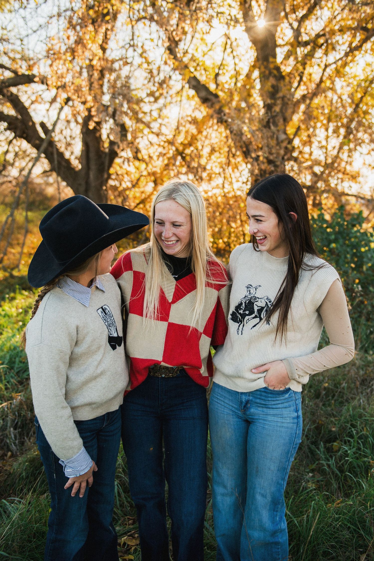 Three women laughing together outdoors in western sweaters with golden autumn trees in the background.