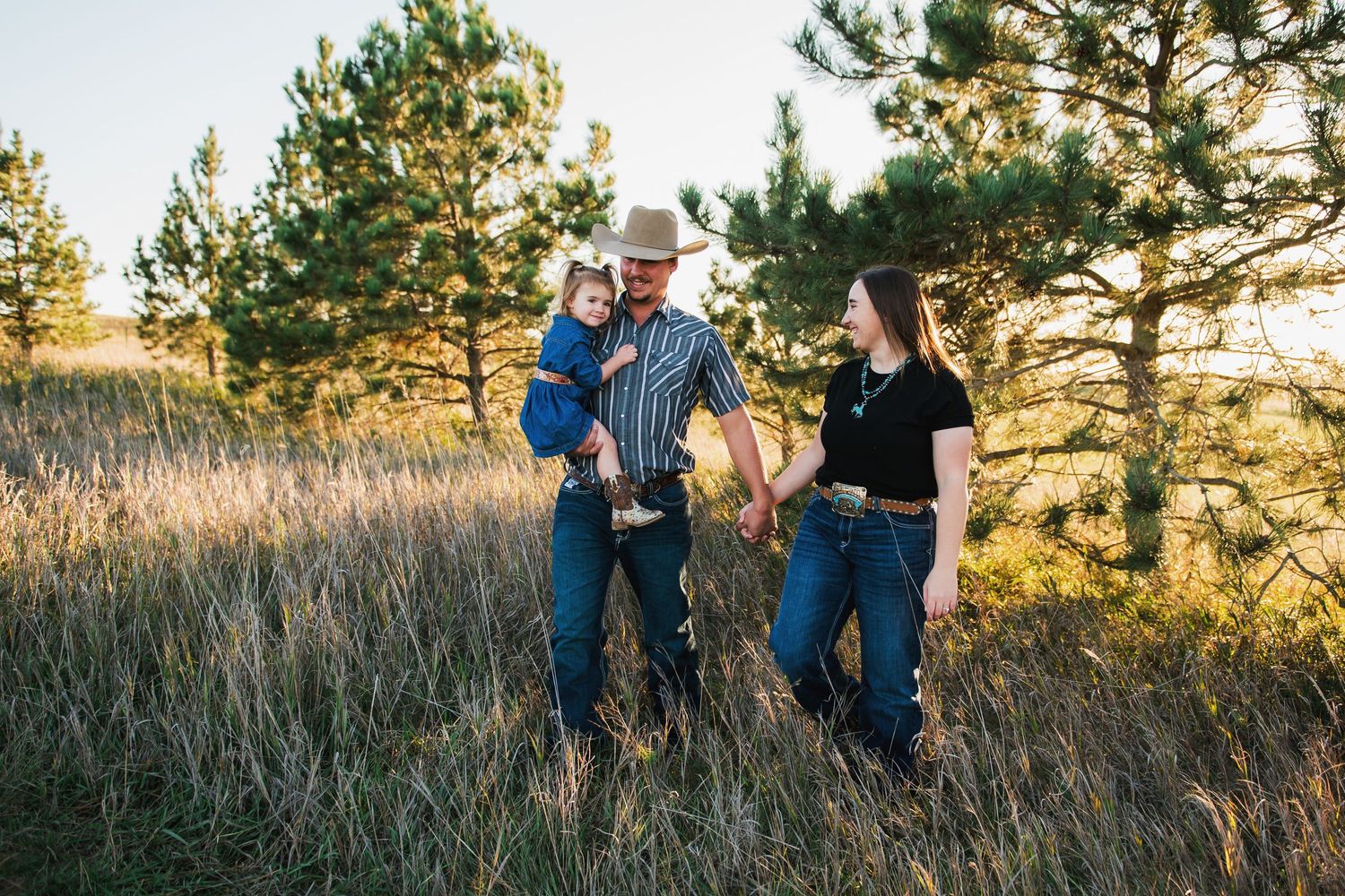 Family of three enjoying golden hour in a meadow surrounded by pine trees, father holding toddler wearing cowboy hat.
