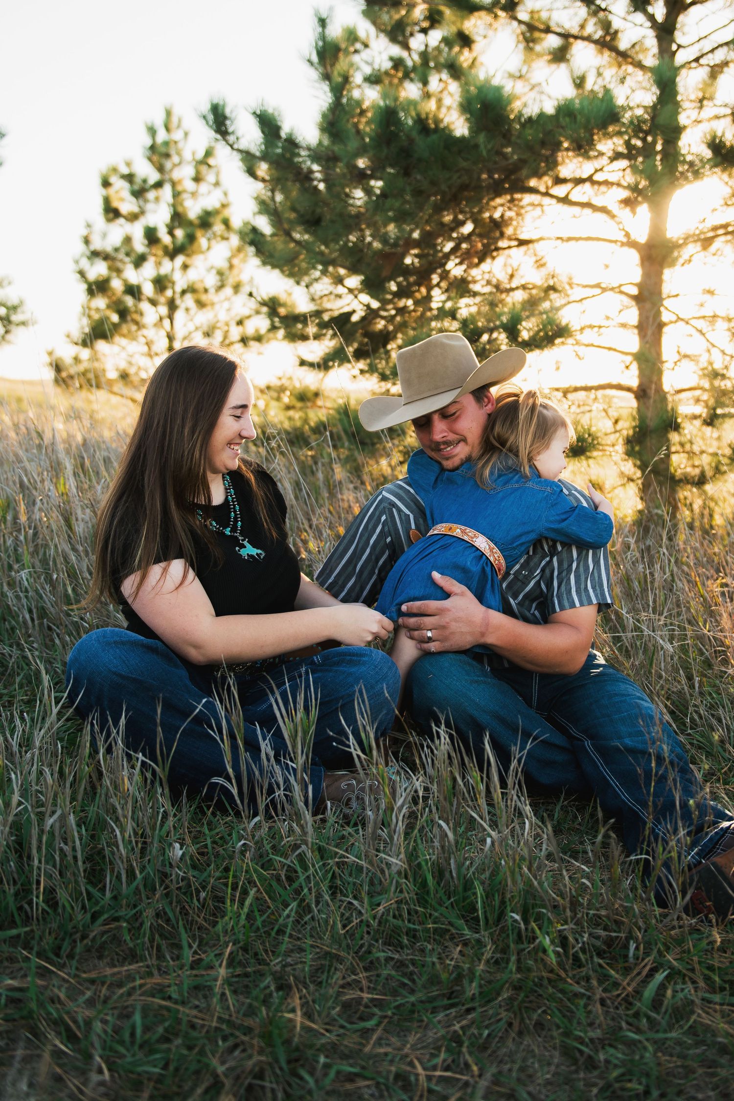 Family of three sitting in tall golden grass at sunset with pine trees in the background.