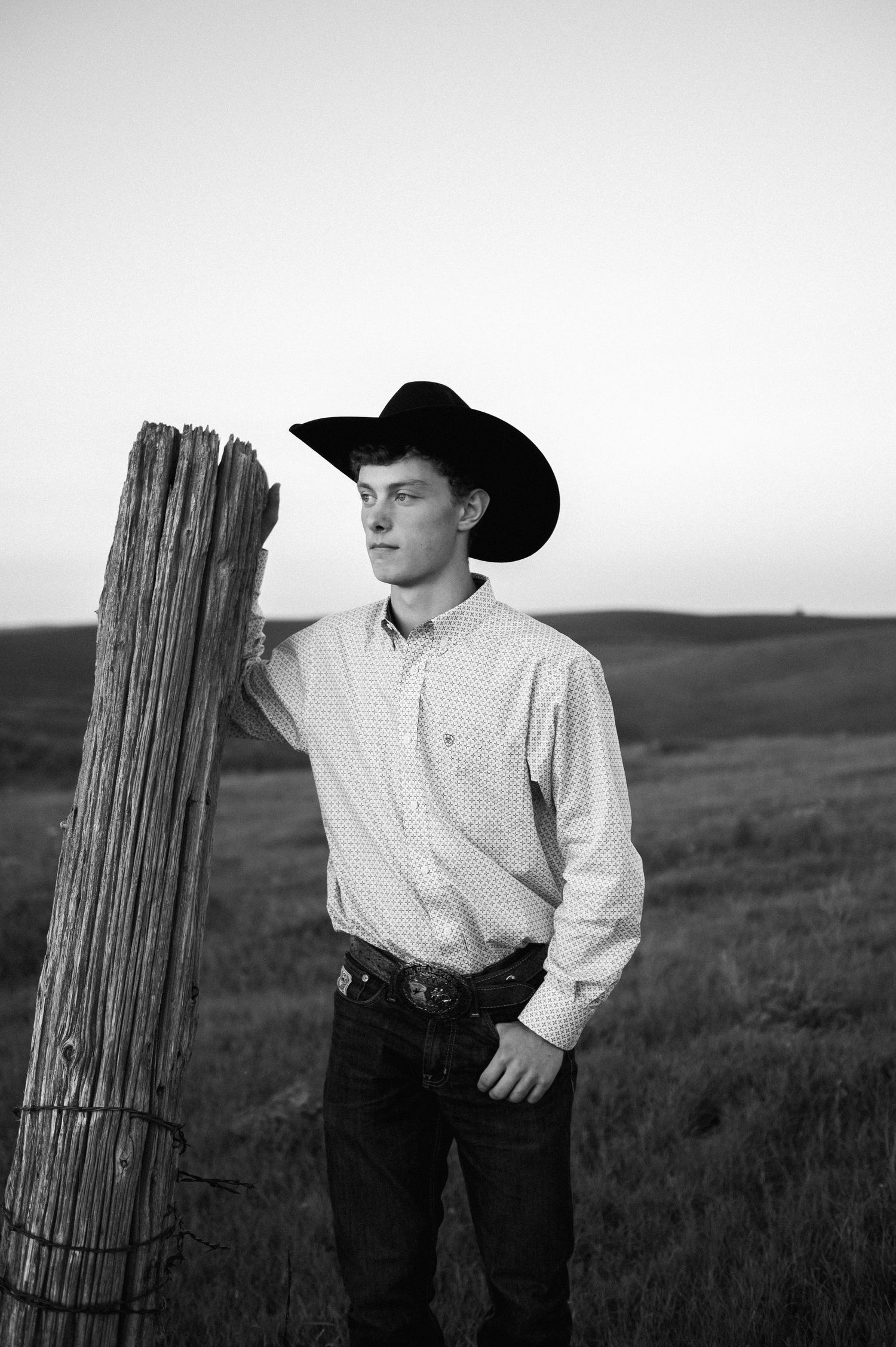 Young cowboy in black hat leaning on a wooden fence post in a black and white prairie photo.