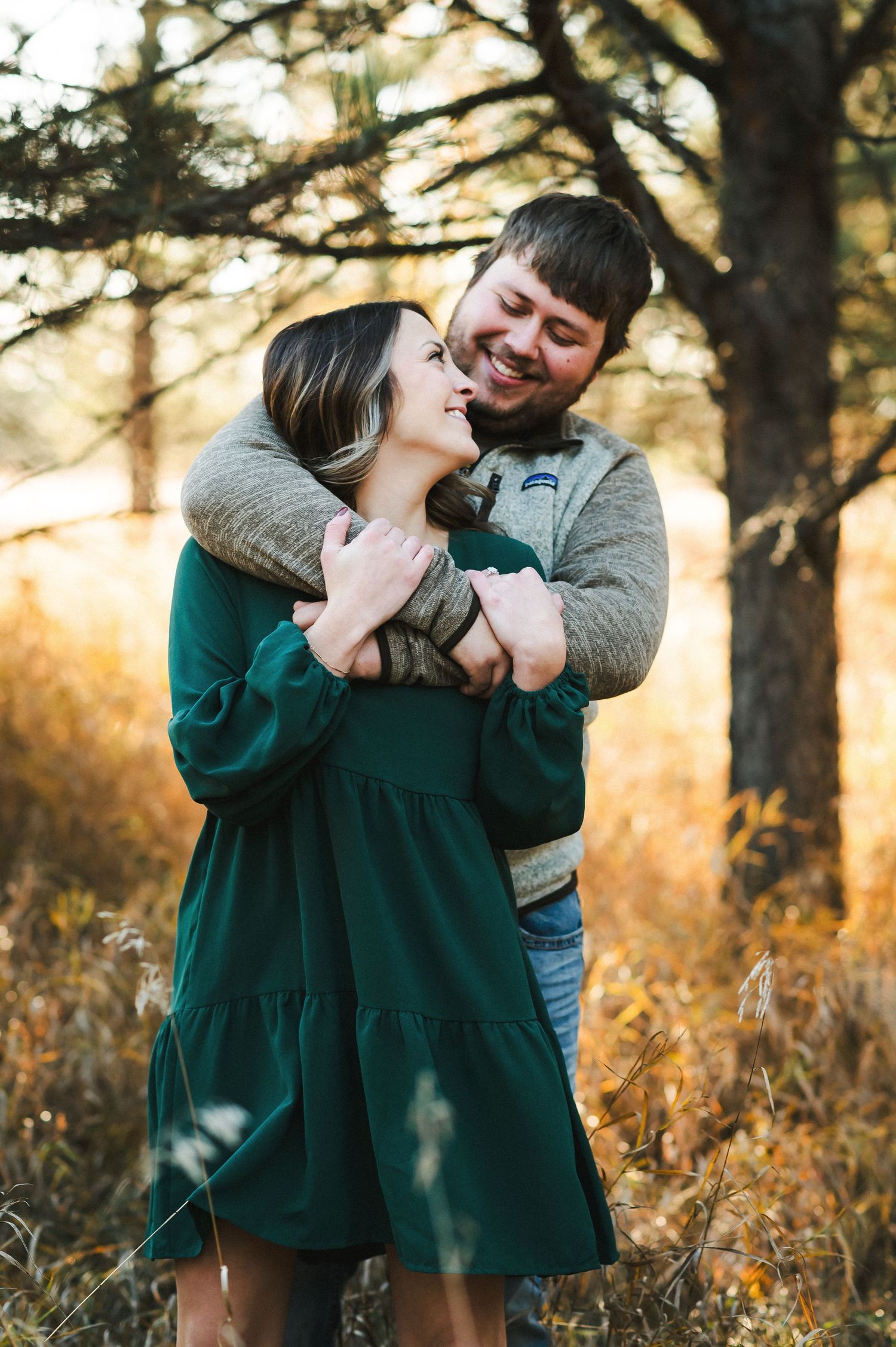 A couple embraces warmly in a golden autumn forest, woman in green dress smiling up at man in gray sweater.