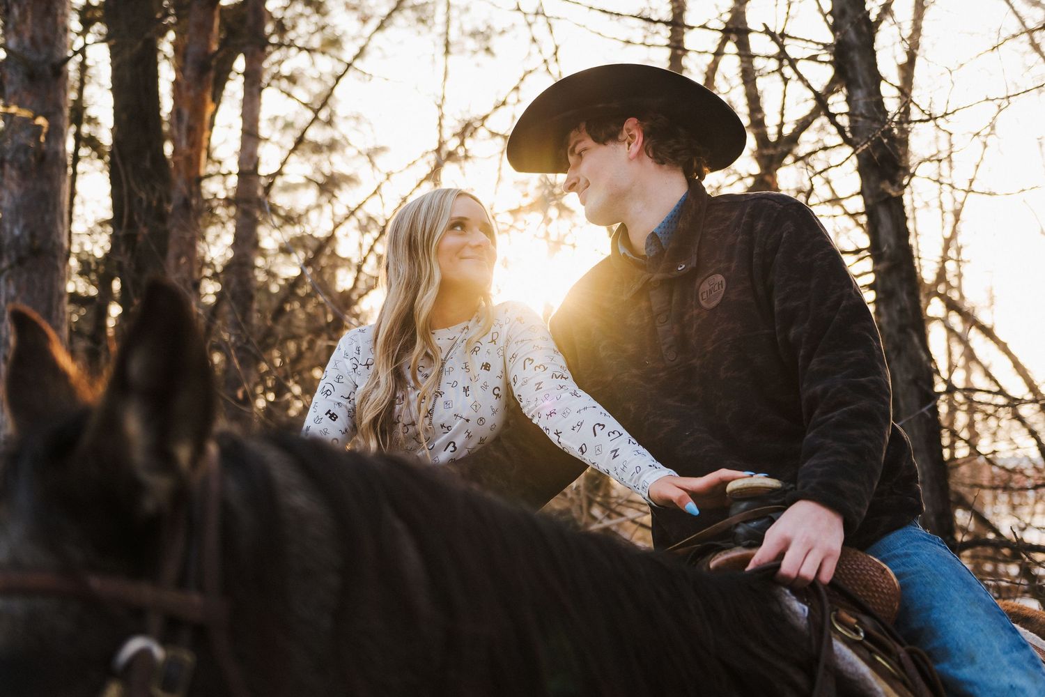 Couple sharing a romantic moment on horseback at golden hour with sun rays filtering through trees.