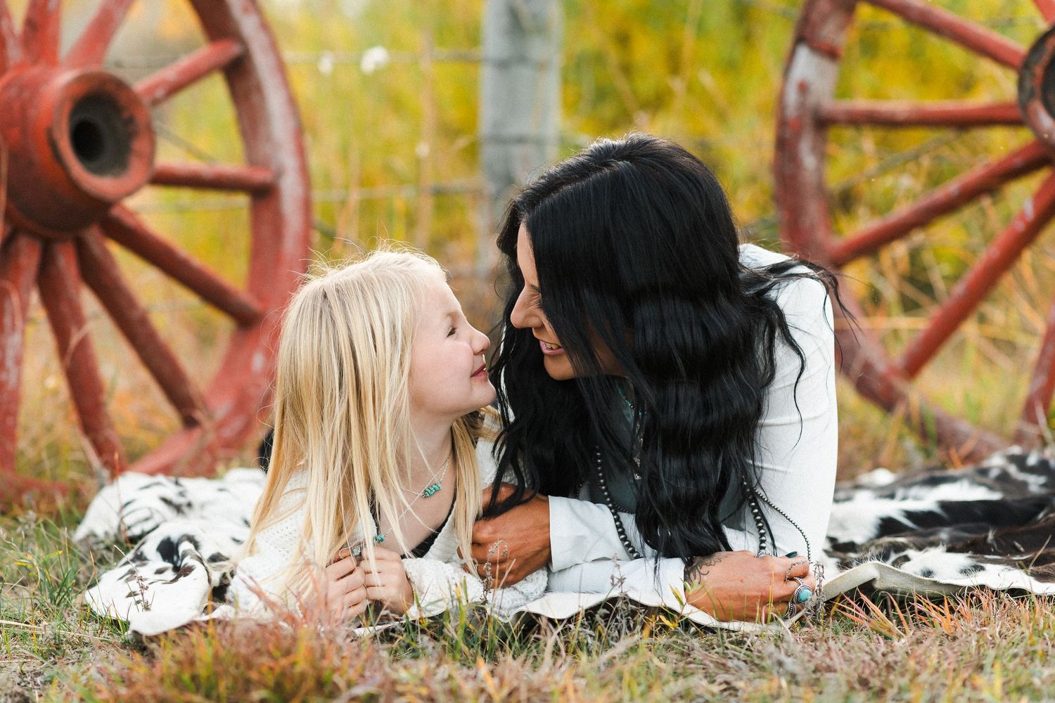 Mother and young daughter lying on a blanket face to face near rustic wagon wheels in autumn.
