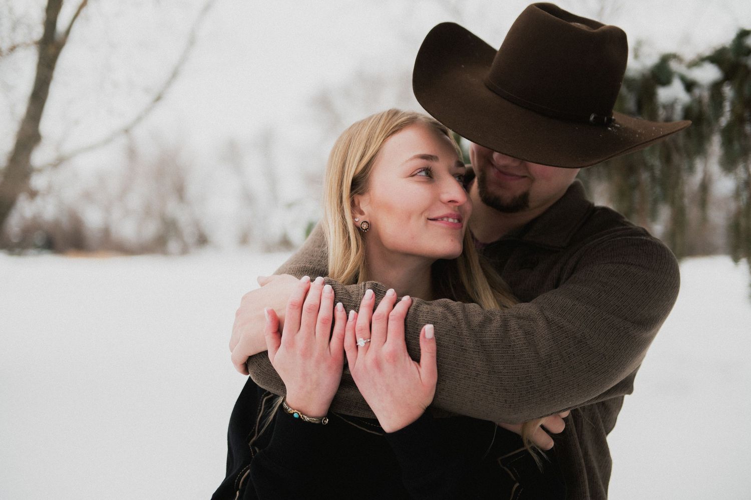 Couple embracing in a snowy winter scene, woman showing engagement ring while man wears a cowboy hat.