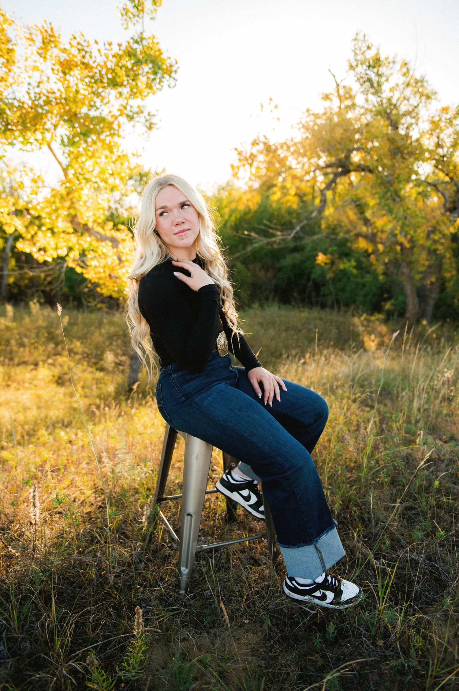 Blonde woman in black top and jeans sits on stool outdoors in golden sunlight surrounded by autumn trees.