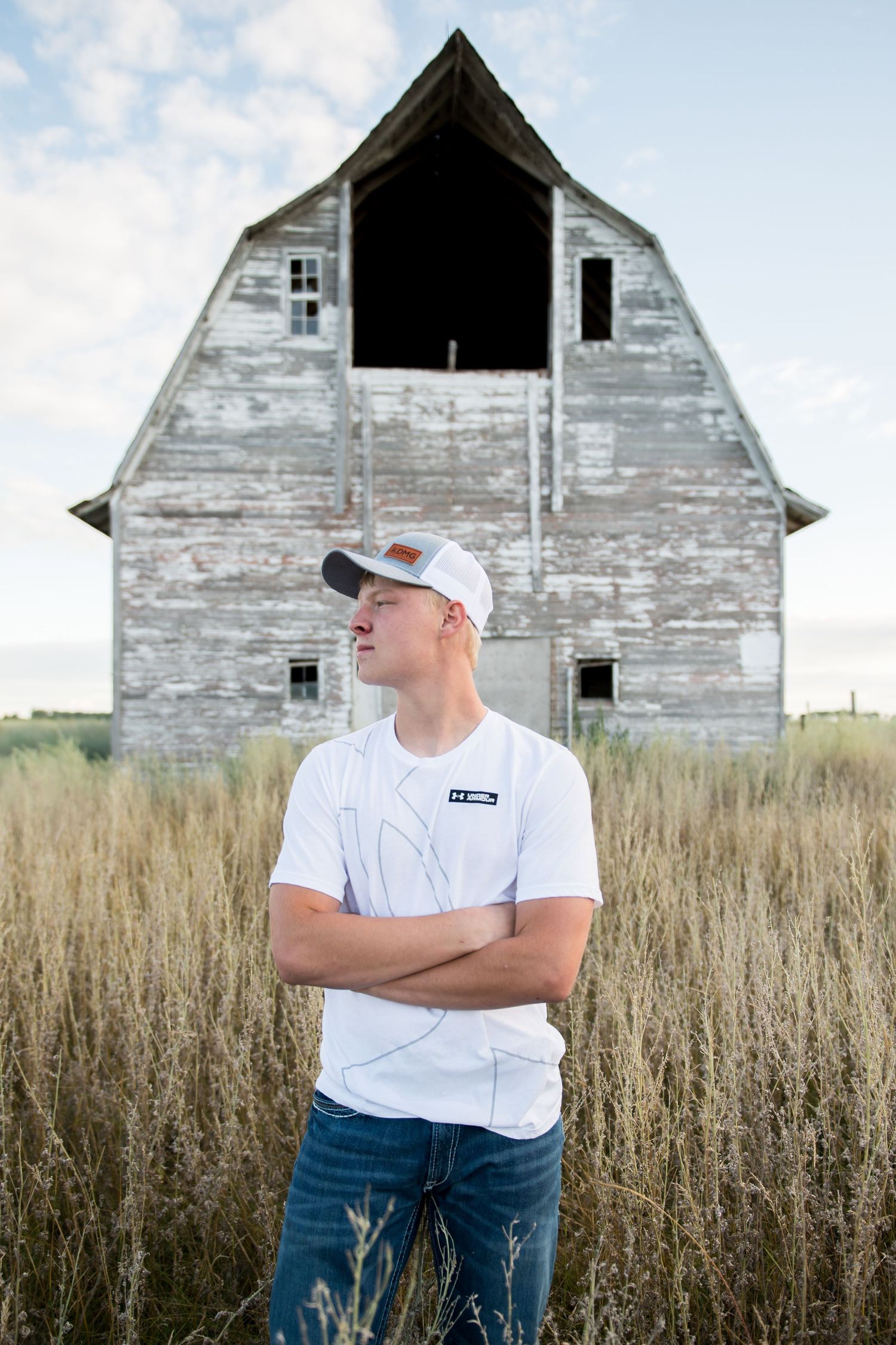Young man in a white shirt and cap standing arms crossed before a weathered white barn in a field.