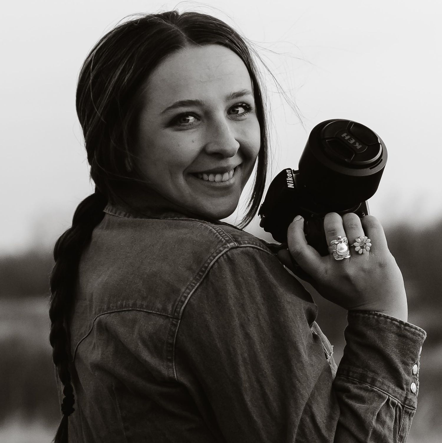 Female photographer smiling over her shoulder holding a camera lens in a black and white portrait.