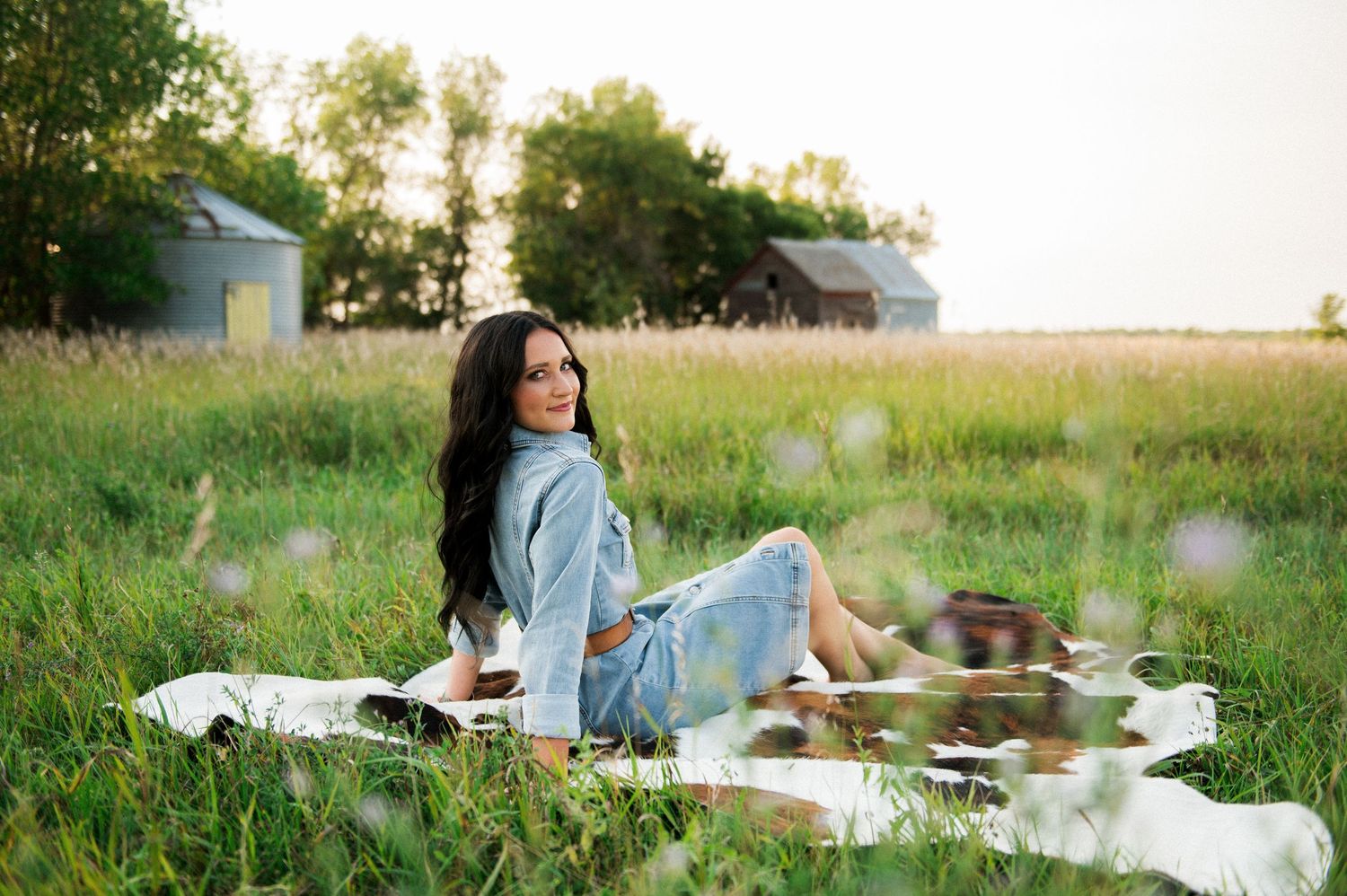 Young woman with long dark hair sits on a blanket in a lush green field near a barn at golden hour.