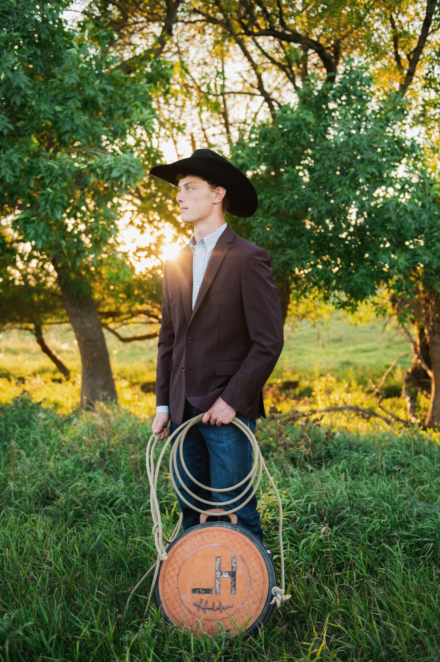 Young man in a black cowboy hat and blazer holding a lasso at golden hour in a green meadow.