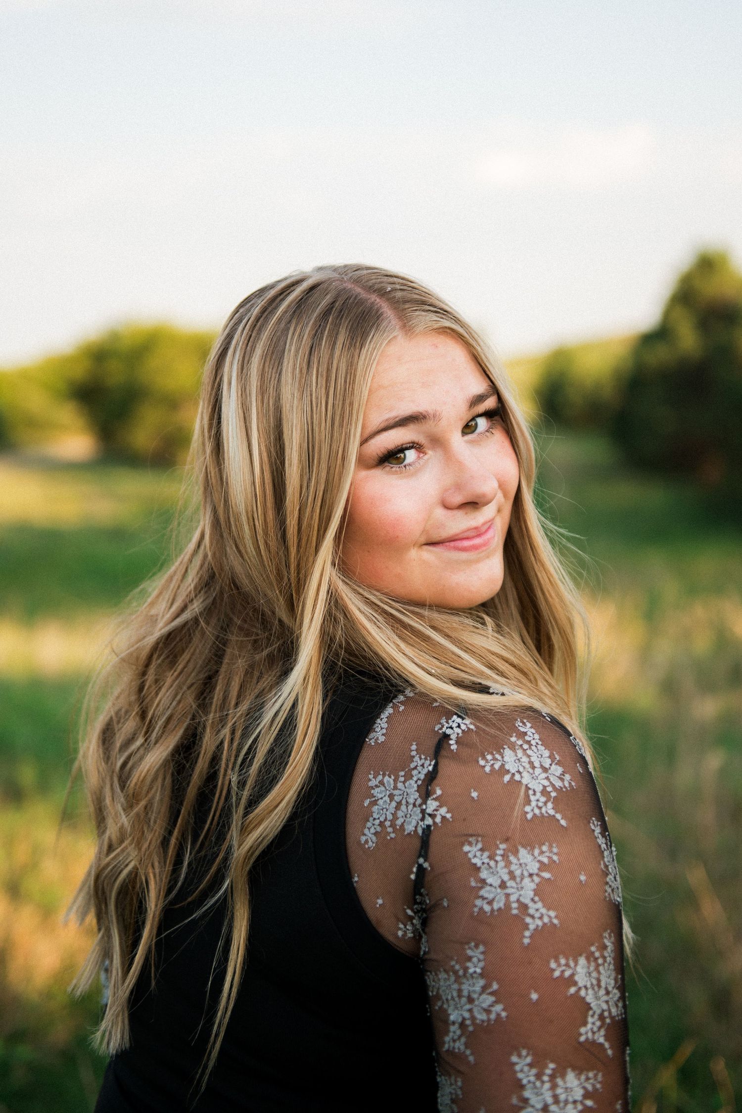 Blonde teenage girl in lace dress looking over her shoulder at golden hour in a green field.
