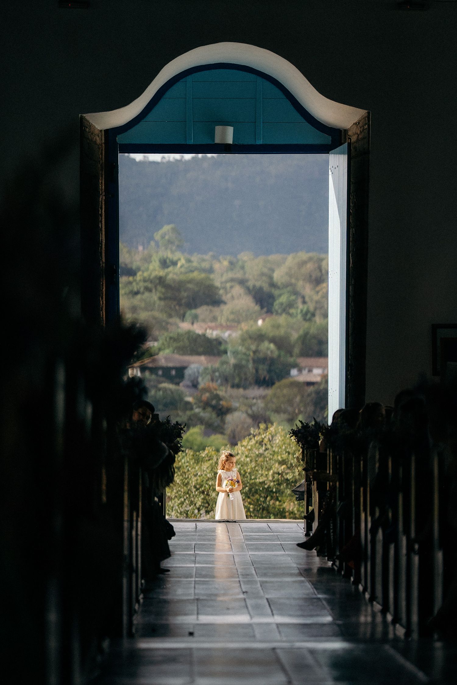 Bride standing in sunlit church doorway with scenic mountain landscape visible beyond the entrance.