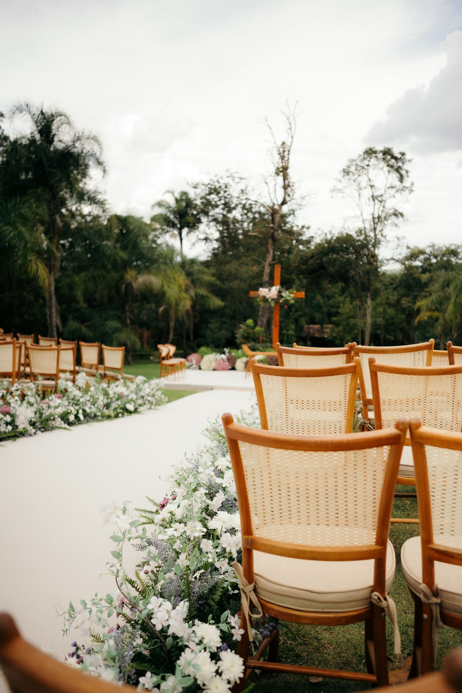 Elegant outdoor wedding ceremony aisle lined with white flowers and wooden rattan chairs leading to an altar surrounded by lush greenery.