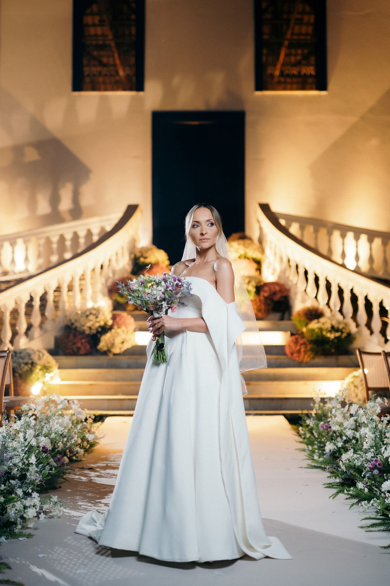 Elegant bride in white off-shoulder gown holds bouquet before illuminated staircase at night wedding venue.