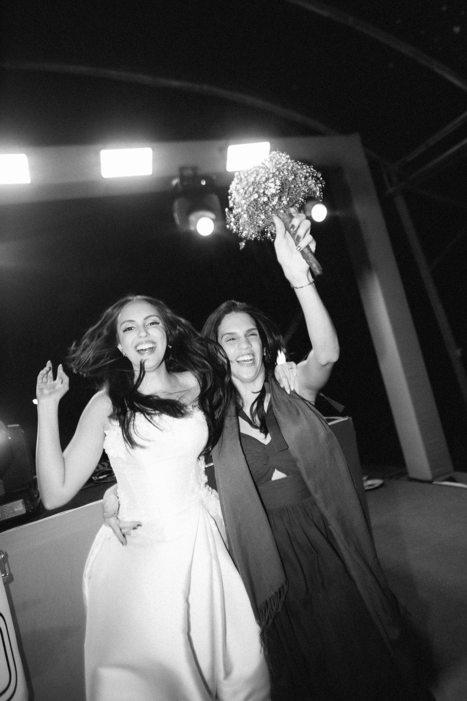 Black and white photo of bride and guest celebrating joyfully with bouquet raised at reception.