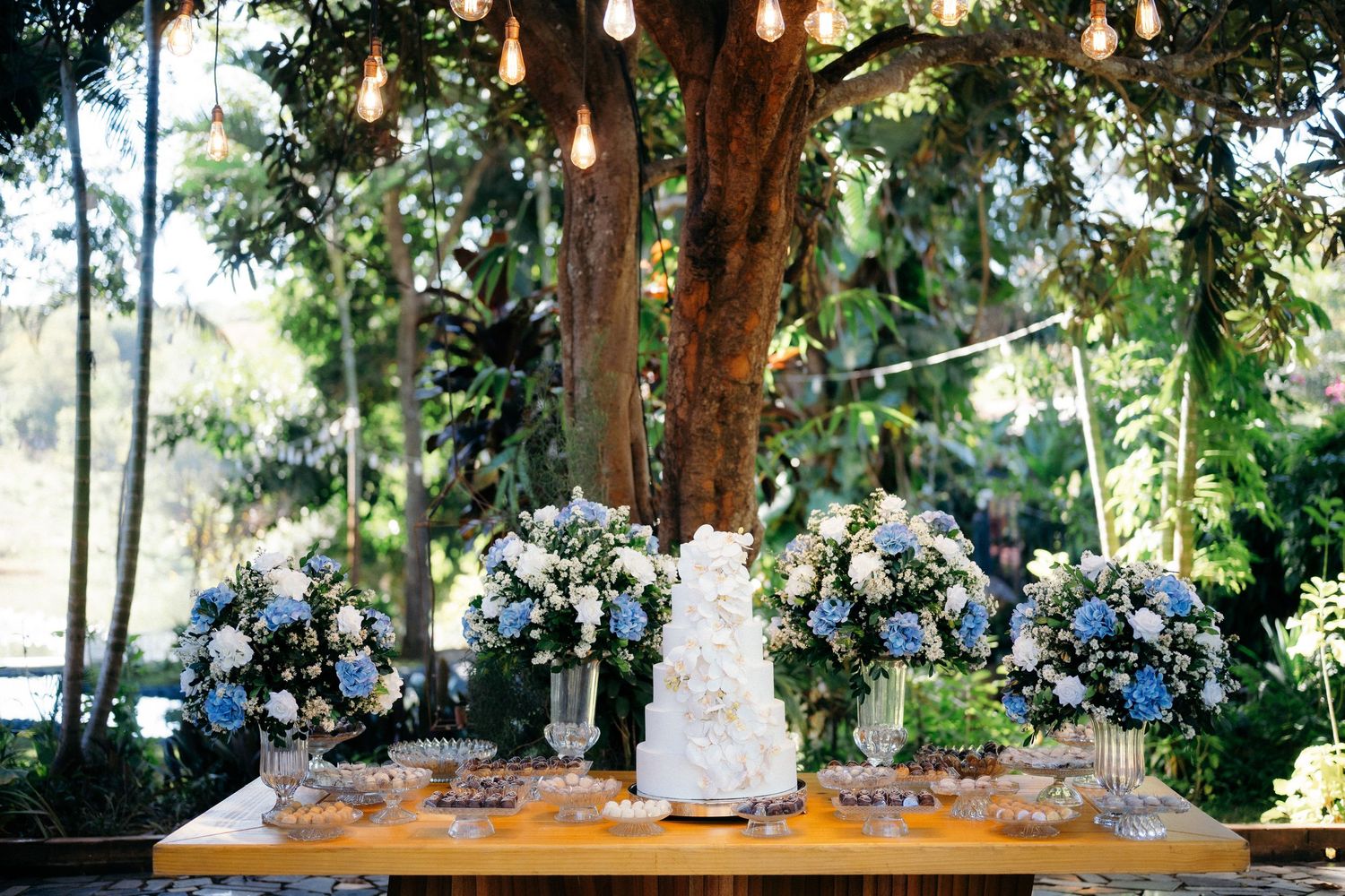 Elegant wedding dessert table with white tiered cake, blue and white floral arrangements under a tree with string lights.