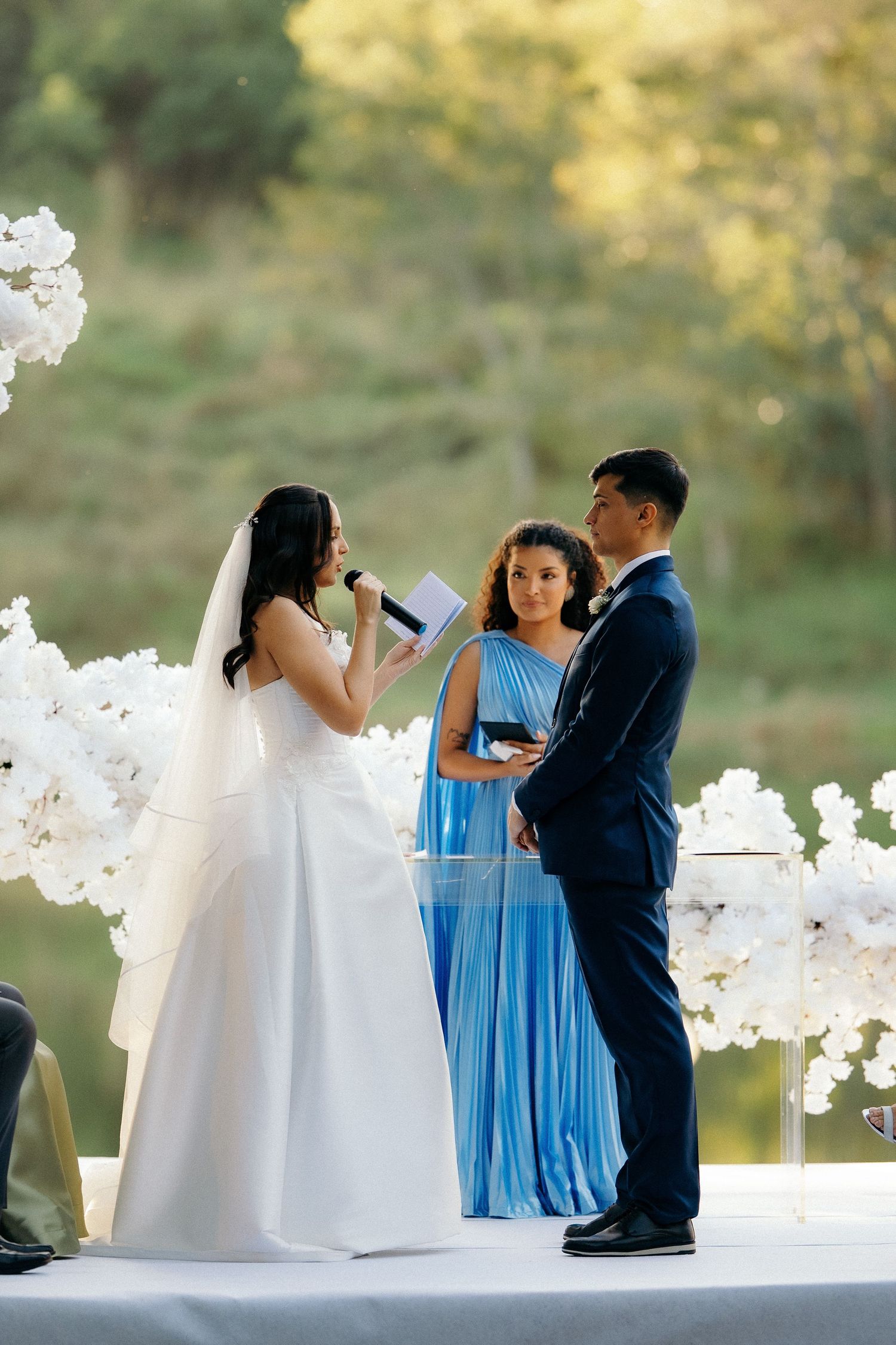 Bride reads vows to groom at outdoor wedding ceremony with white floral arrangements and female officiant.