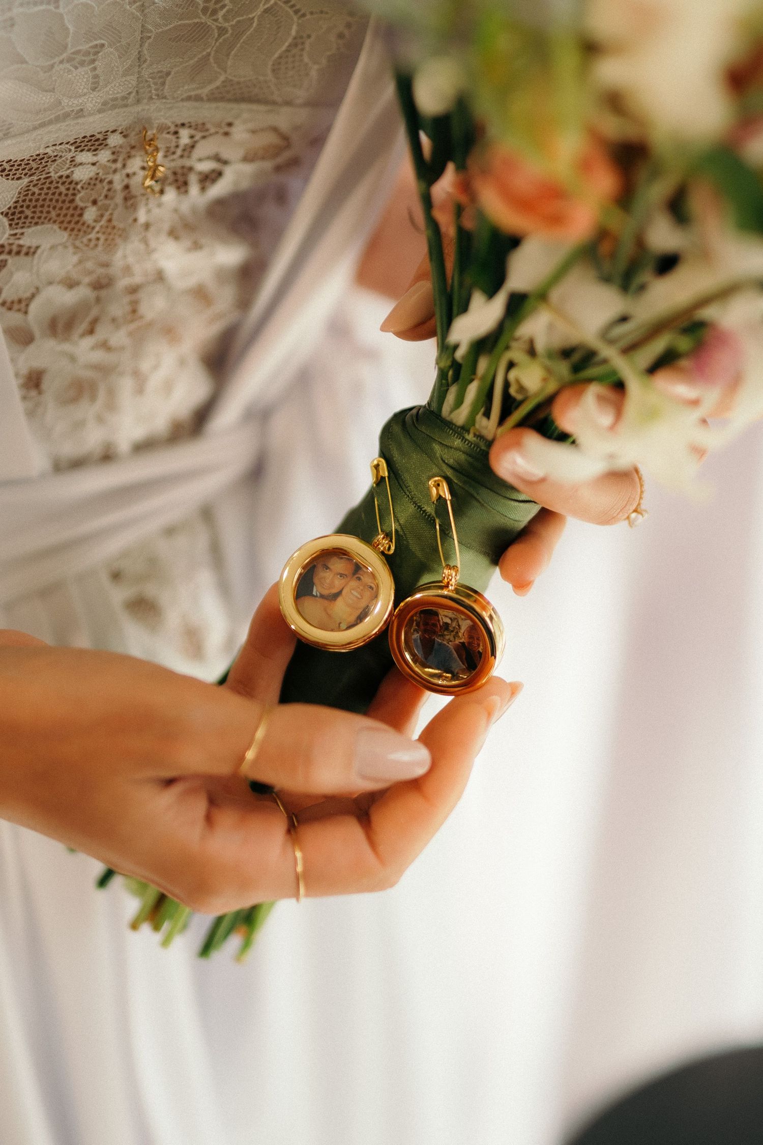 Bride holding wedding bouquet with gold locket charms containing photos, wearing lace dress and gold ring.