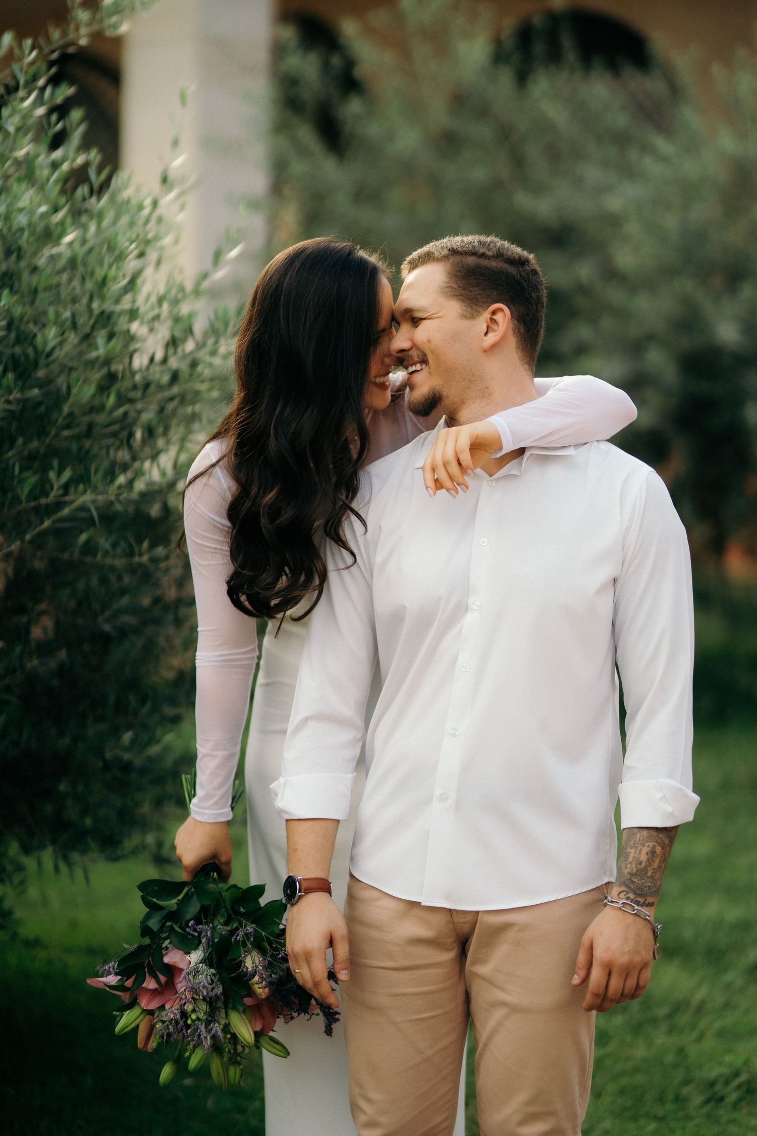 Smiling couple embracing outdoors, woman in white dress holding bouquet, man in white shirt with tattoo.