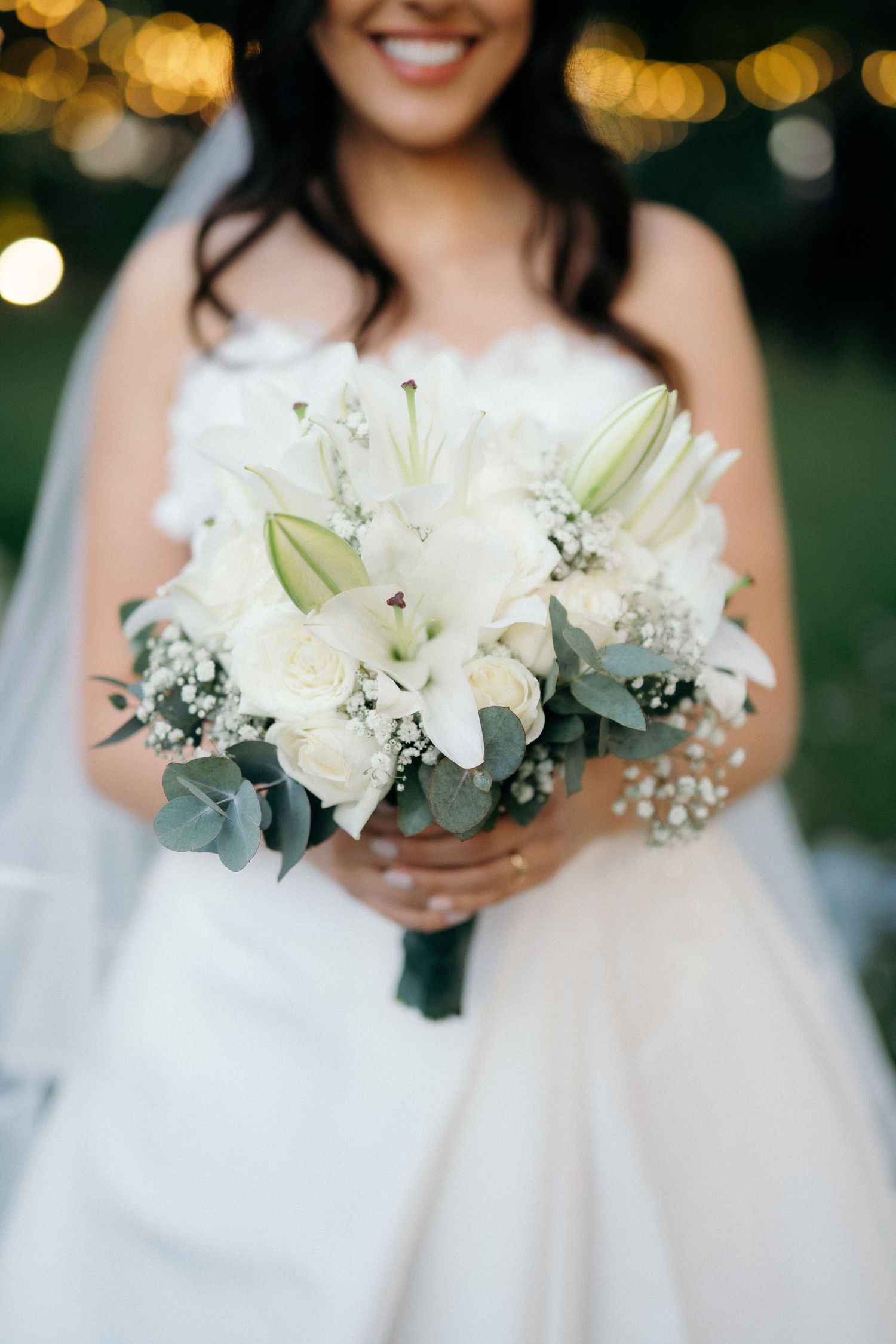 Bride holding elegant white bouquet of lilies, roses, and eucalyptus at outdoor wedding.