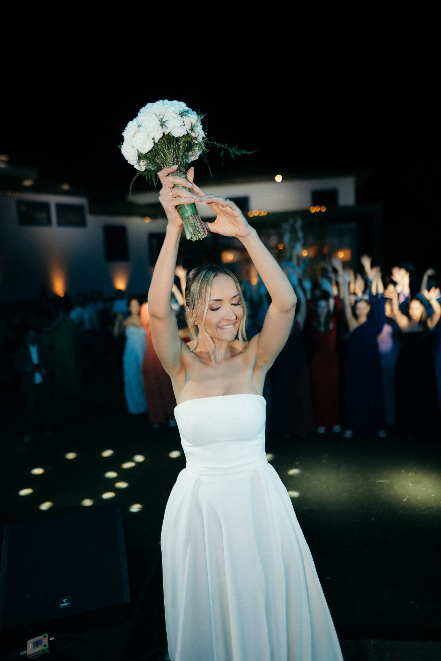 Bride in white strapless gown raises bouquet overhead smiling before bouquet toss at evening reception.