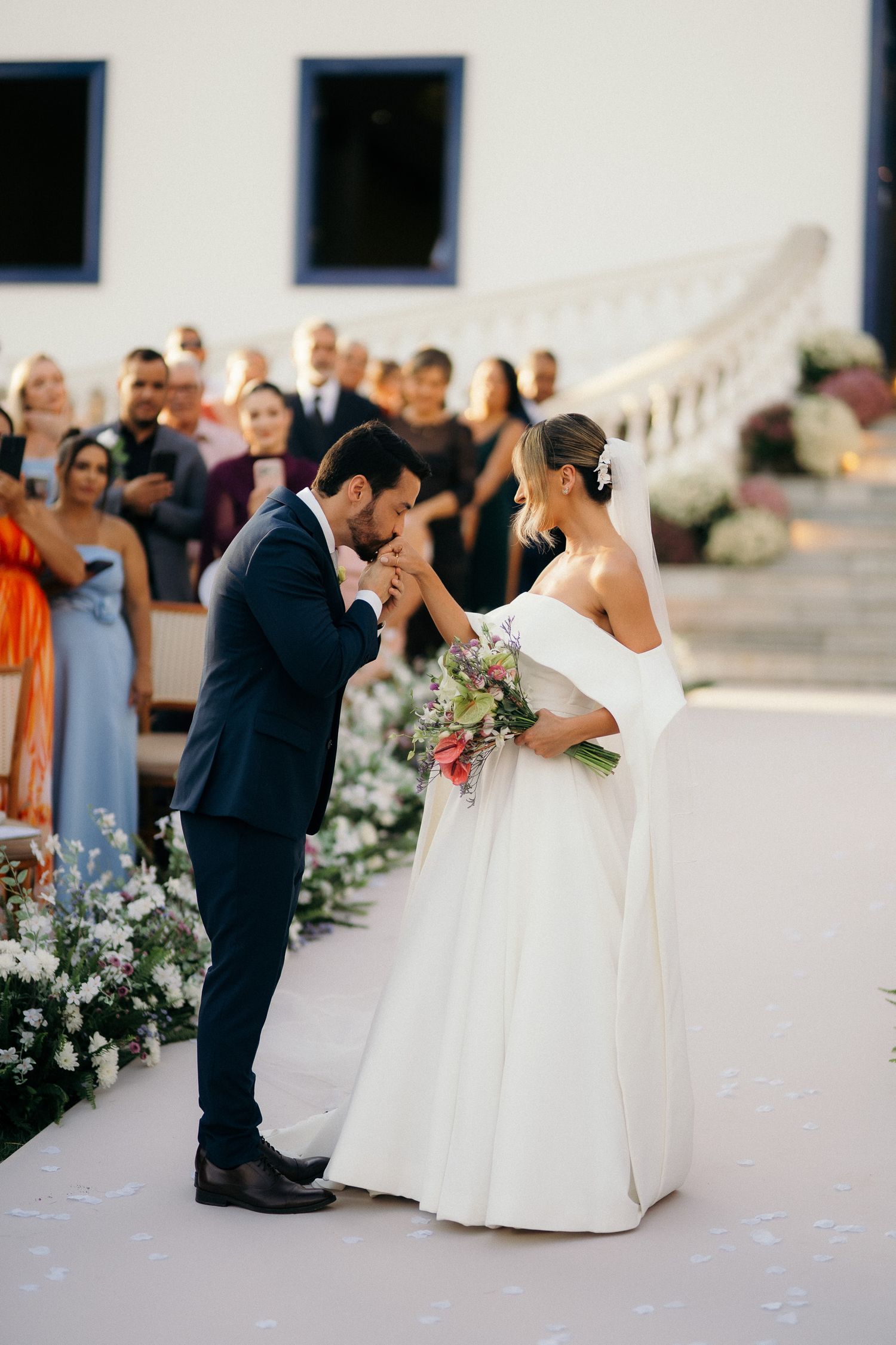 Groom in navy suit kisses bride's hand during outdoor wedding ceremony while guests watch.