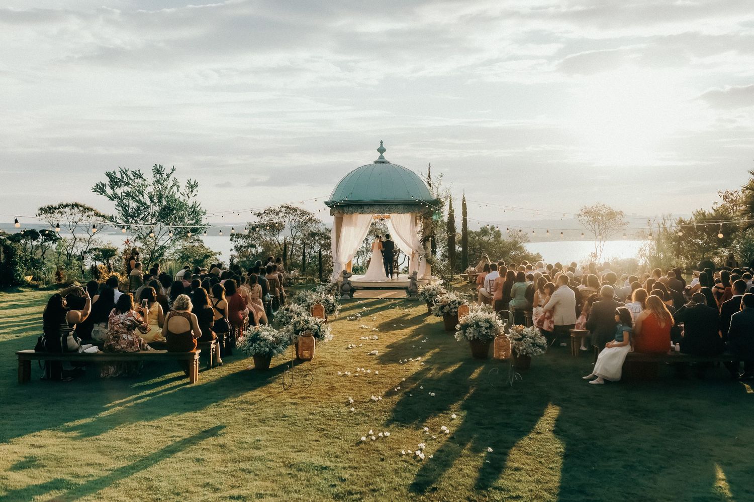 Outdoor wedding ceremony at sunset with guests seated on a lawn around a blue gazebo altar surrounded by floral arrangements.