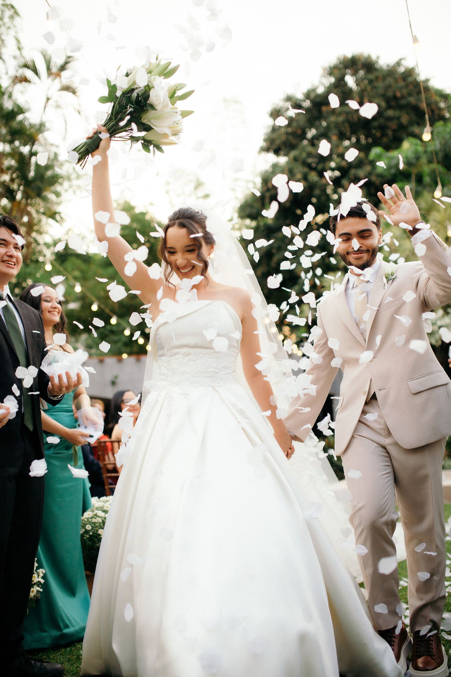 Joyful newlyweds walking down aisle as guests toss white flower petals in celebration.