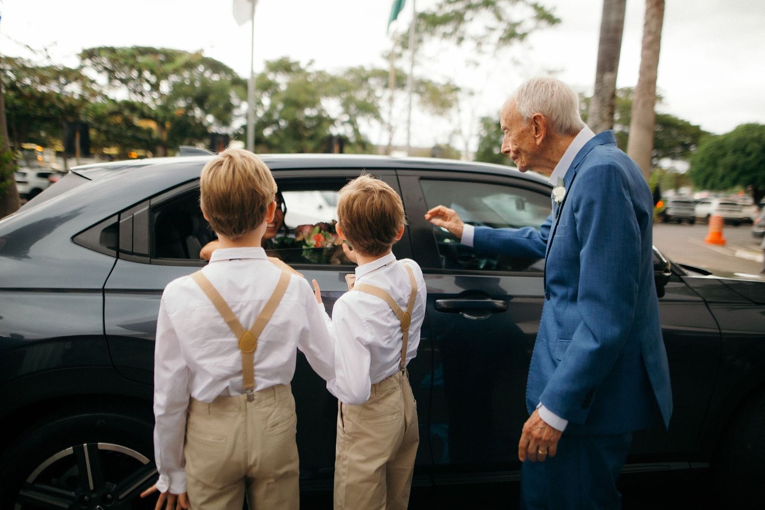 Elderly man in blue suit showing a black car to two young boys in white shirts and suspenders.