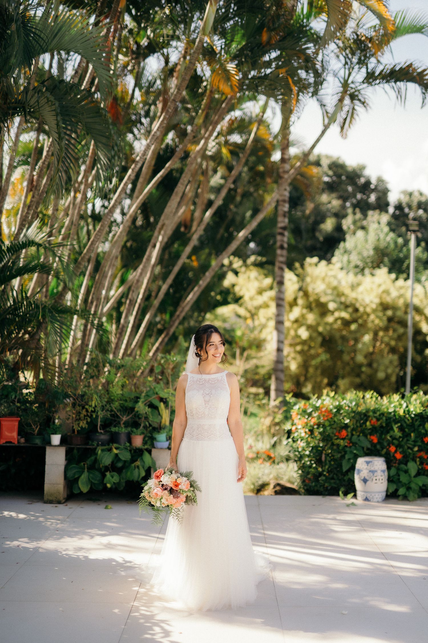 Smiling bride in white tulle gown holding colorful bouquet outdoors among tropical palm trees.