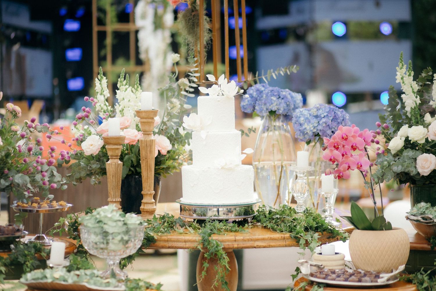 Elegant white tiered wedding cake surrounded by colorful floral arrangements on a decorated reception table.