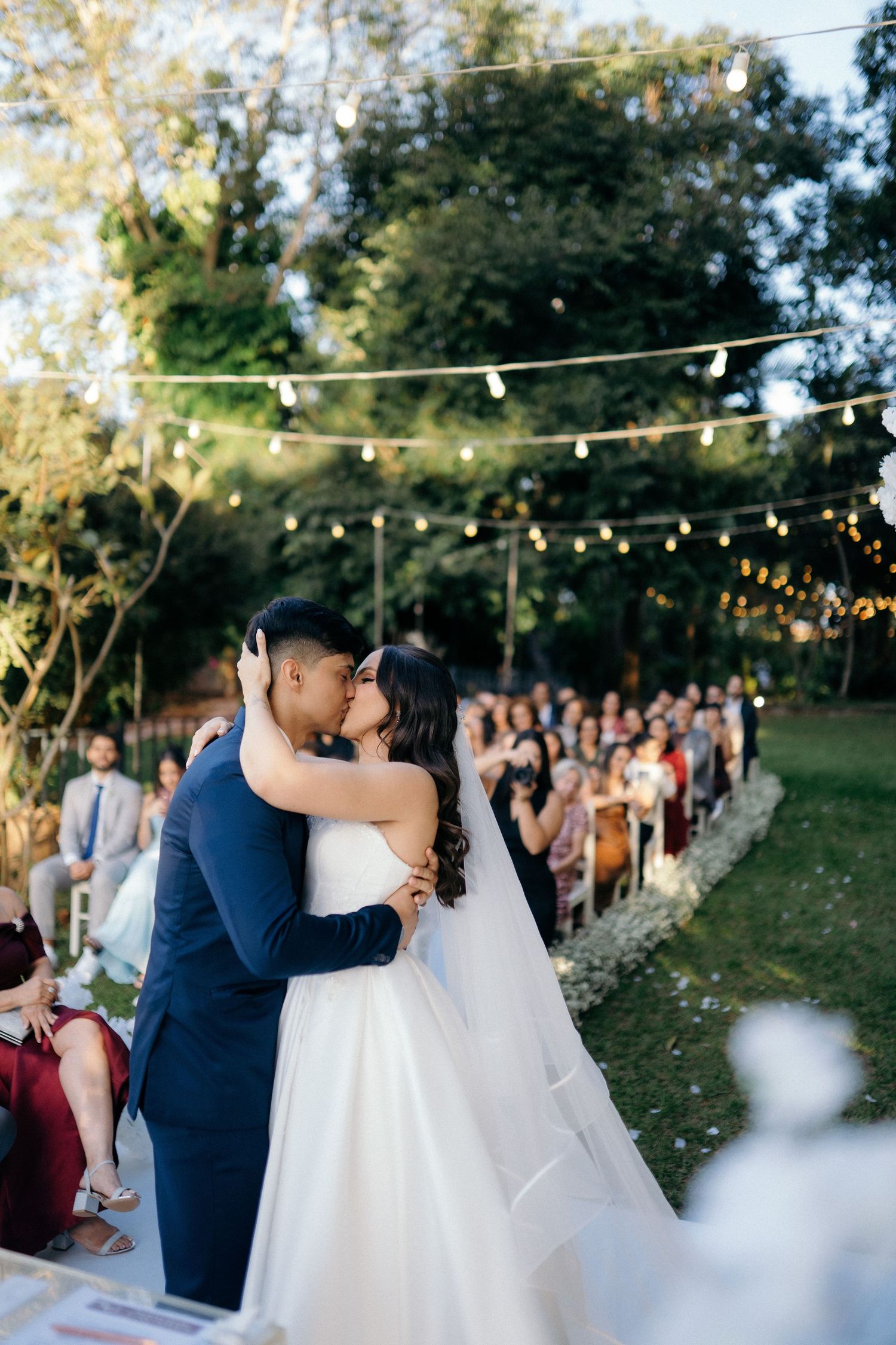 Bride and groom share first kiss at outdoor wedding ceremony with string lights and seated guests.