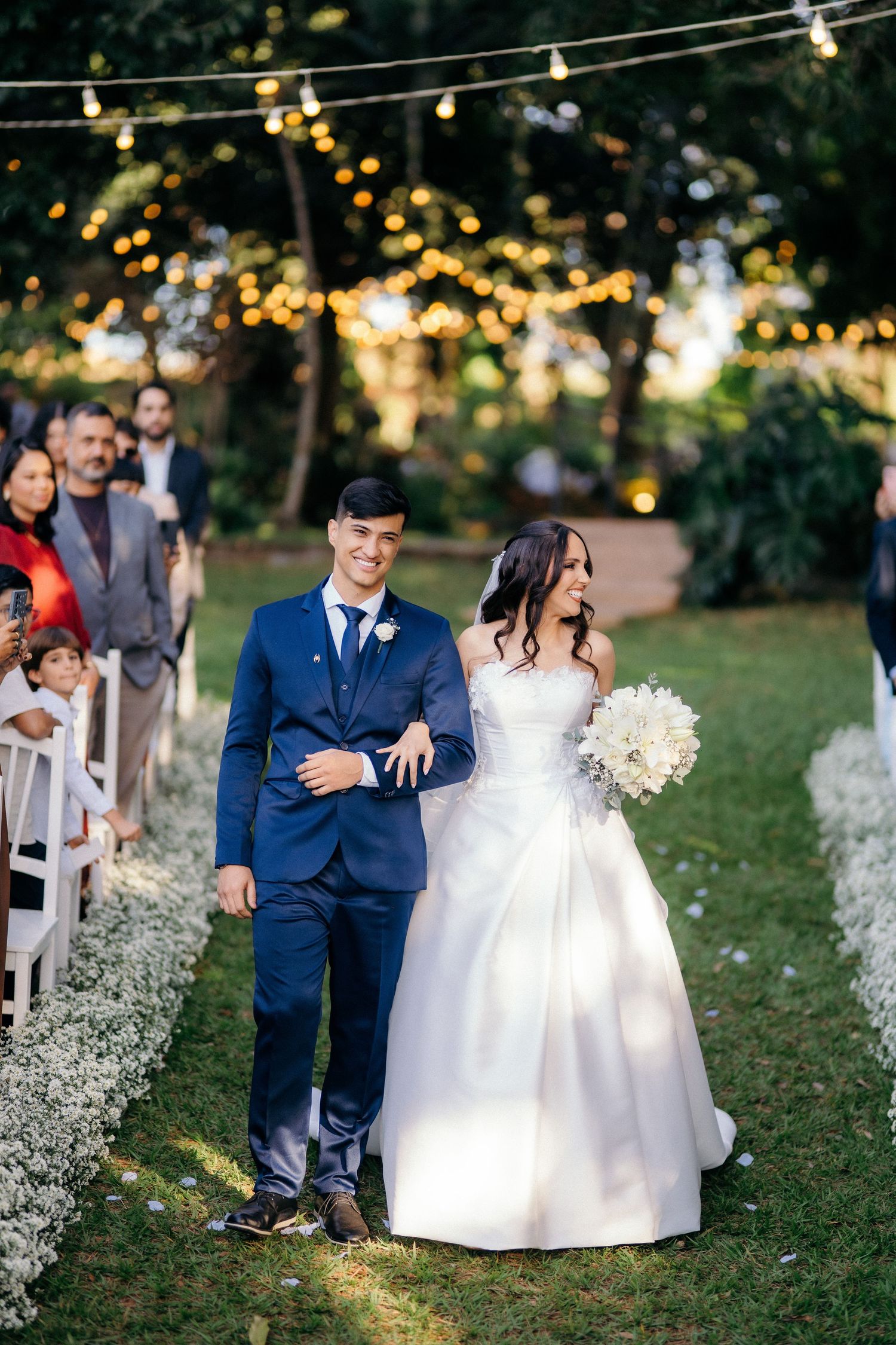 Bride and groom walking down outdoor aisle under string lights surrounded by wedding guests.