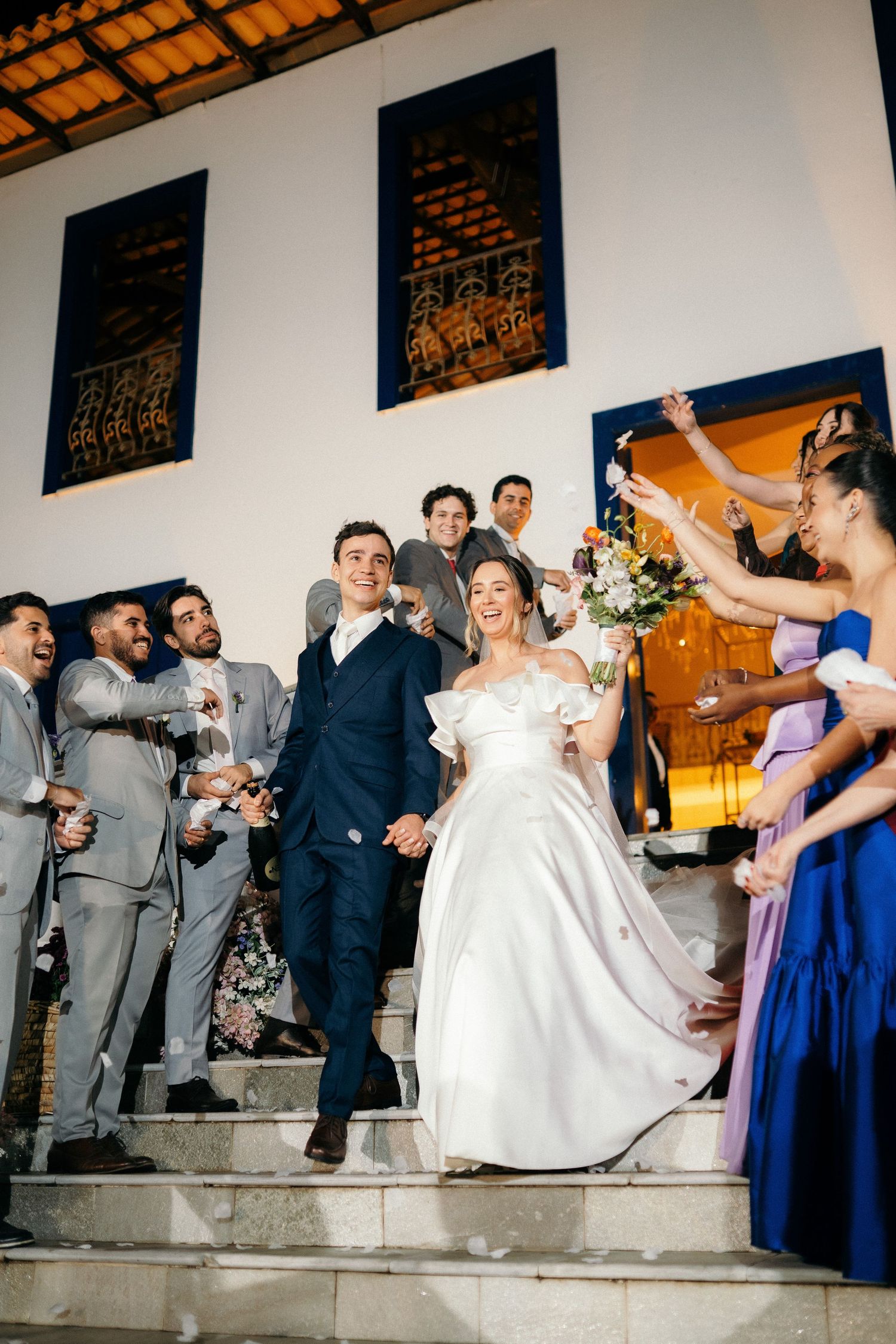 Bride and groom exit church ceremony on steps, surrounded by cheering wedding party in blue and gray attire.