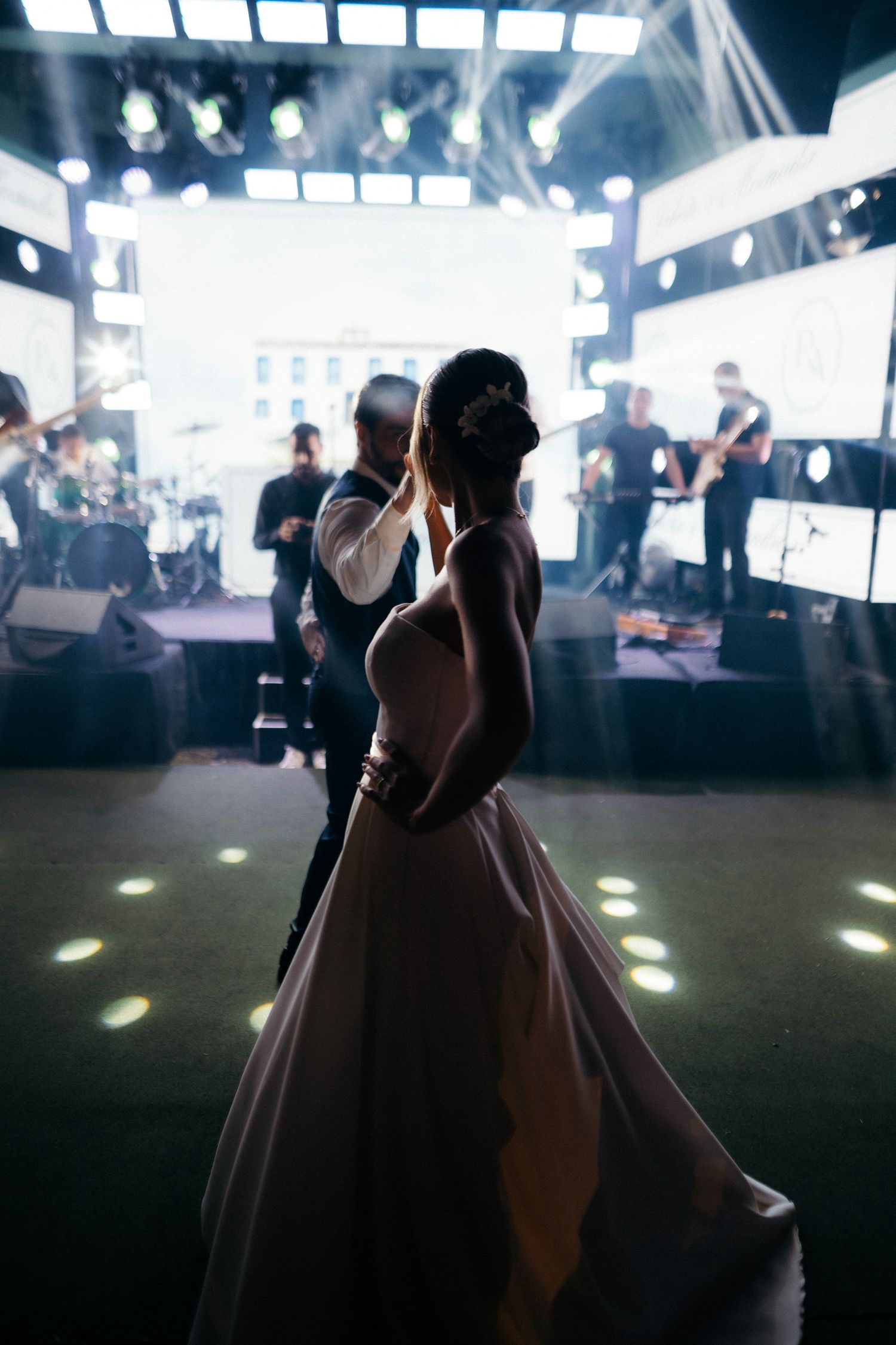 Bride and groom share first dance in silhouette before a live band on a lit wedding stage.