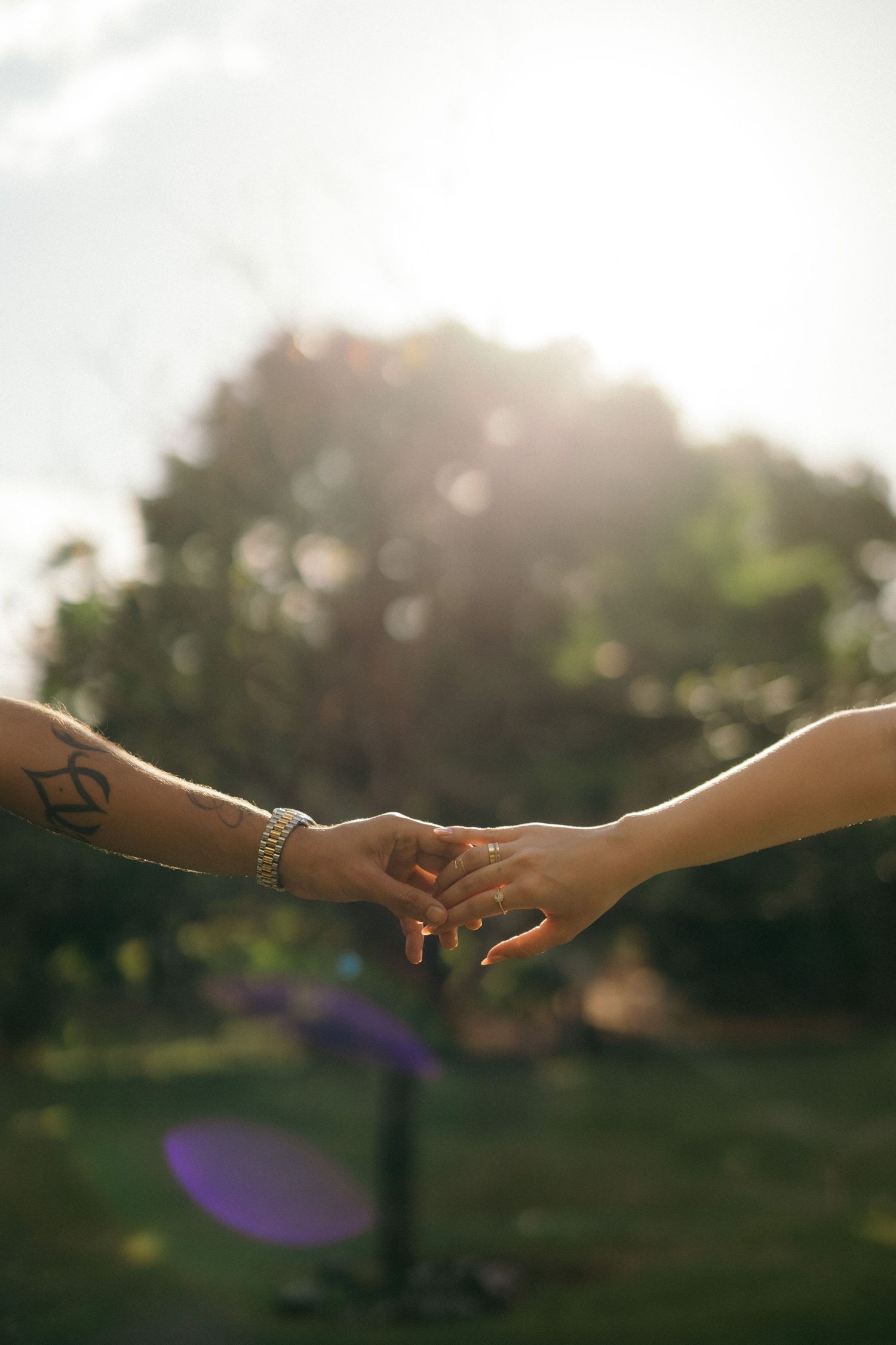 Two hands gently touching outdoors in sunlight, one with a tattoo and bracelet, the other with a ring, against a green bokeh background.