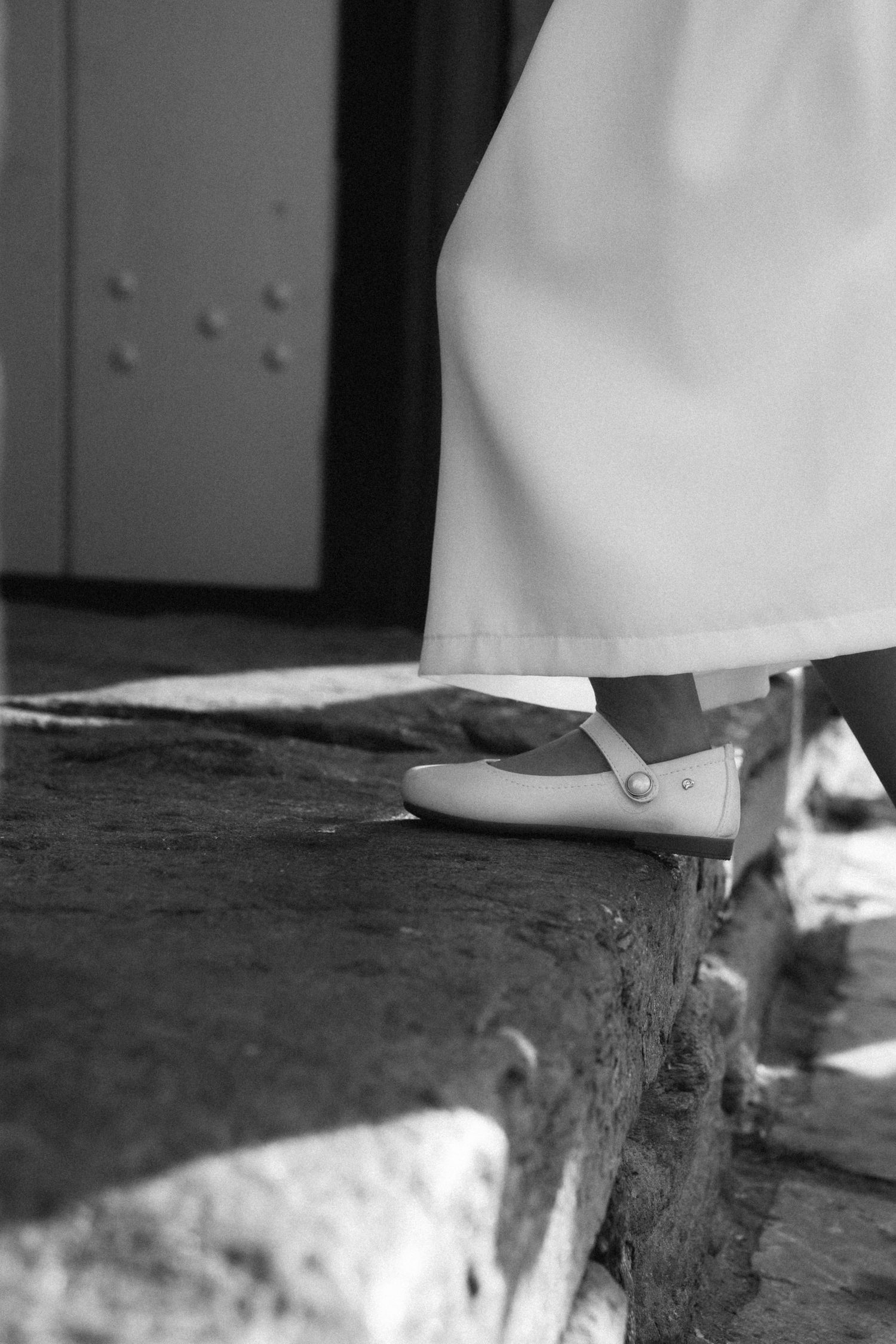 Close-up black and white photo of bride's white flat shoe and wedding dress hem on stone steps.