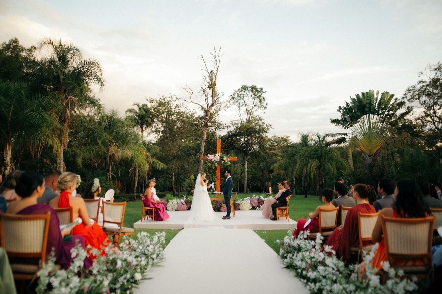 Elegant outdoor wedding ceremony with white aisle, floral arrangements, and guests seated among tropical palm trees.