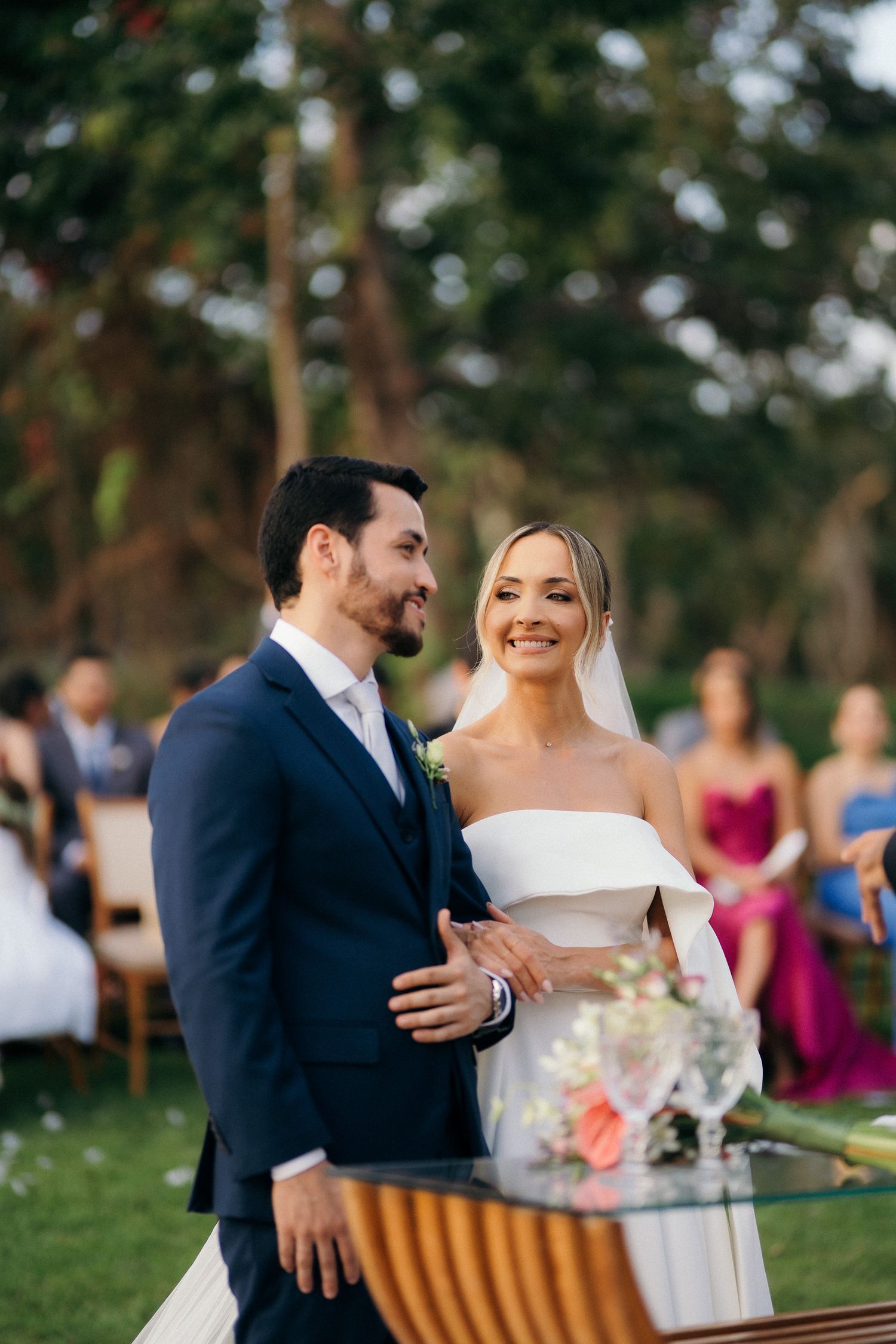 Smiling bride in white off-shoulder gown and groom in navy suit stand together at outdoor wedding ceremony.
