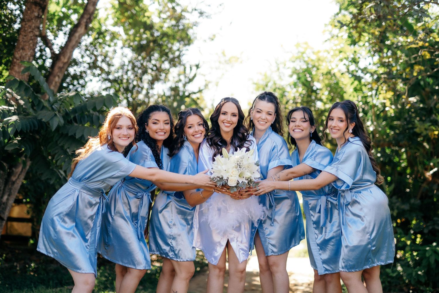 Bride in white robe holds bouquet surrounded by six bridesmaids in matching blue satin robes outdoors.
