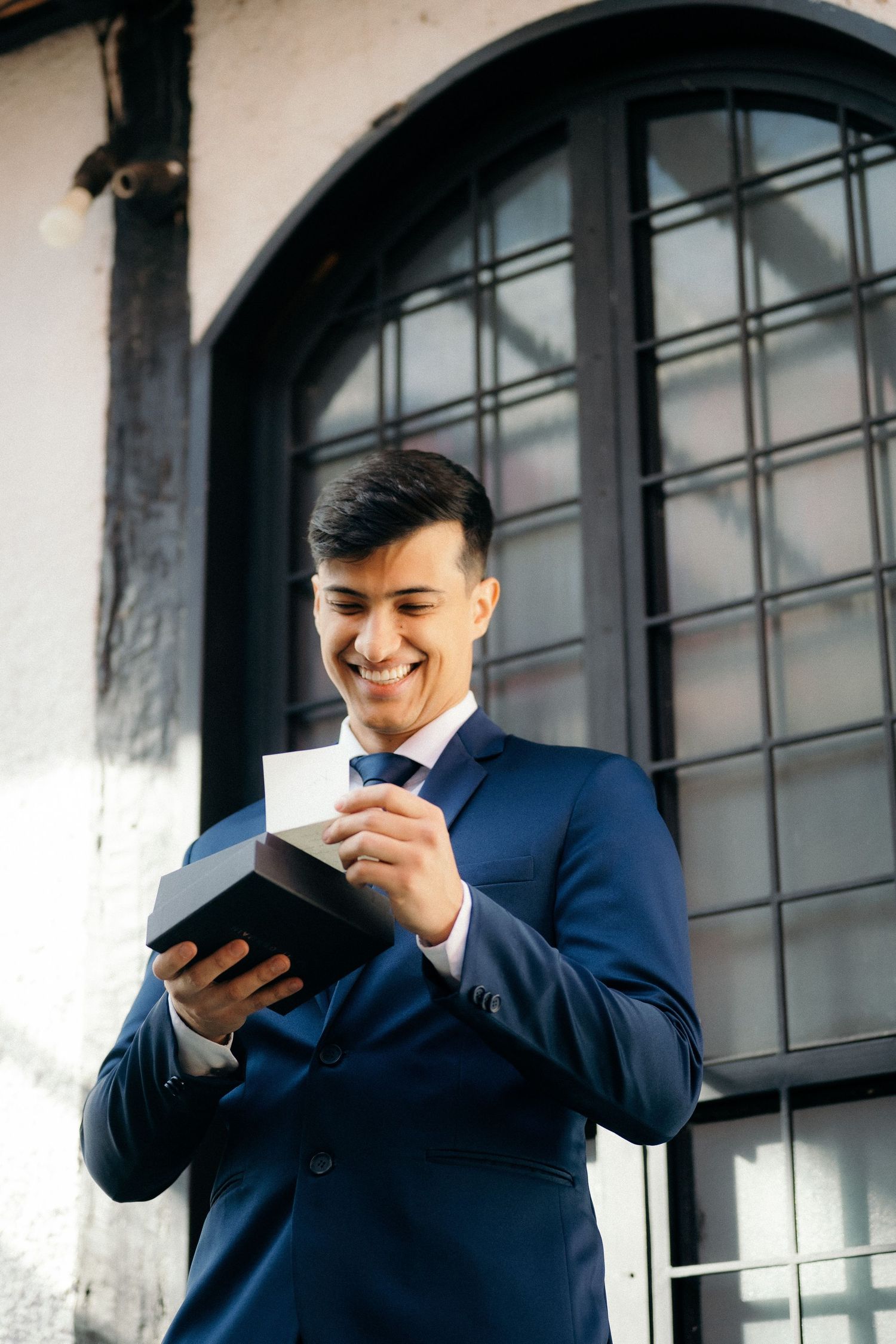 Handsome groom in a navy blue suit smiling while opening a gift box outside a building.