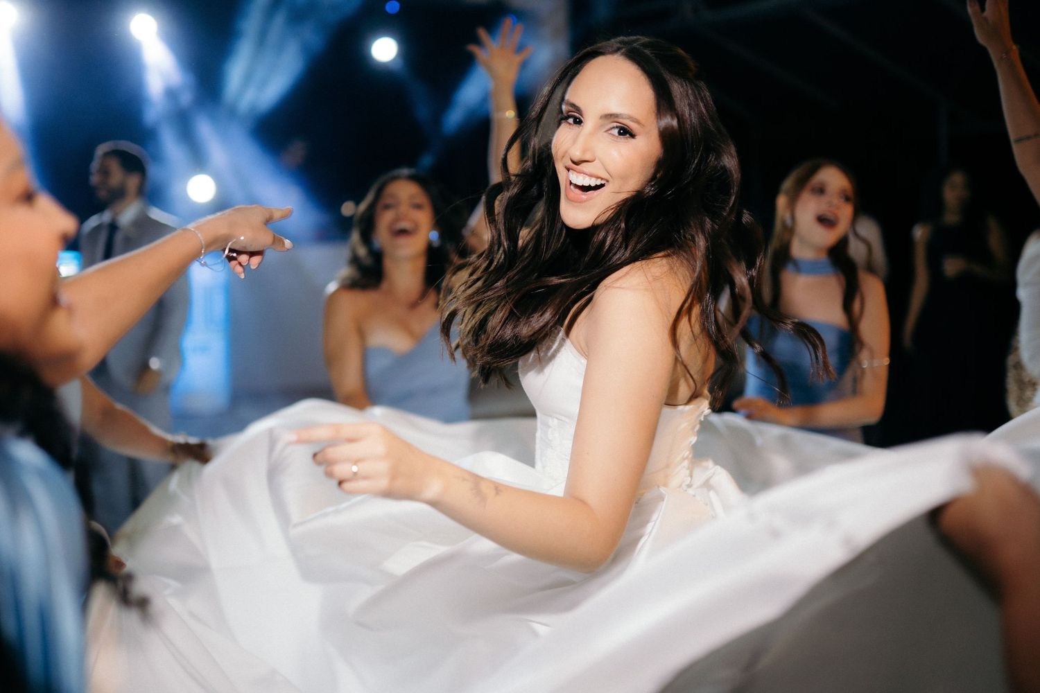 Joyful bride in white strapless gown laughs and dances with bridesmaids in blue at wedding reception.
